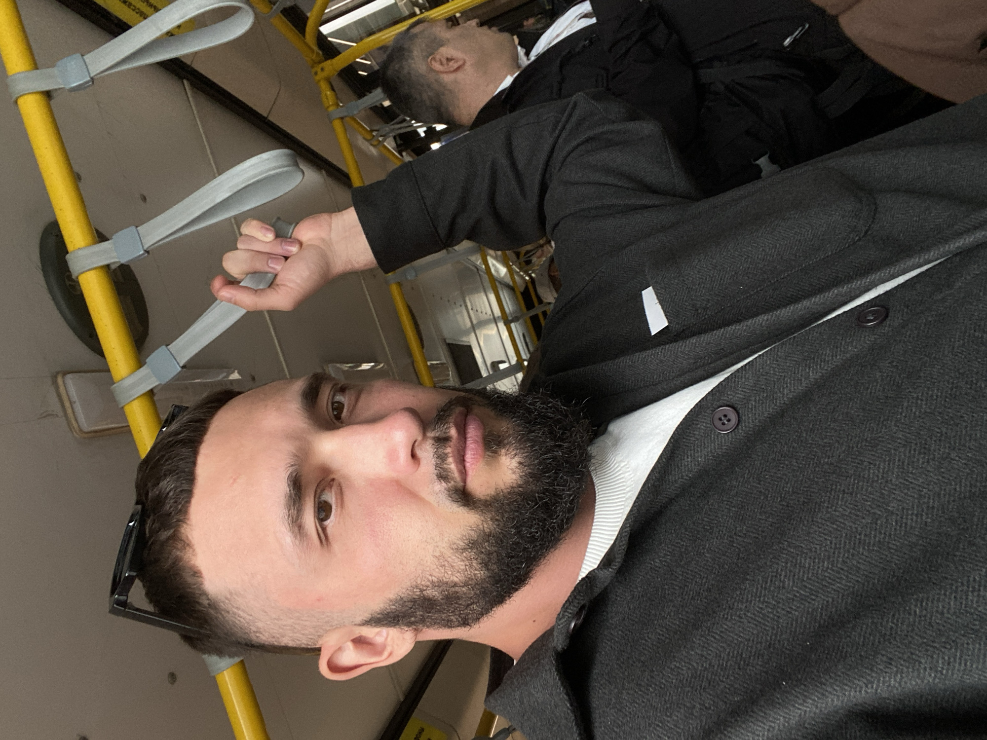 A man with a beard and short hair is taking a selfie on a bus. He is wearing a dark grey jacket over a white shirt. He is holding onto a grey strap attached to a yellow pole. Another man in a black jacket and white shirt is visible in the background, seated on the bus. The bus appears to be in motion, indicated by the strap the man is holding. The location is the city of Novoye Martyanovo, Russia.