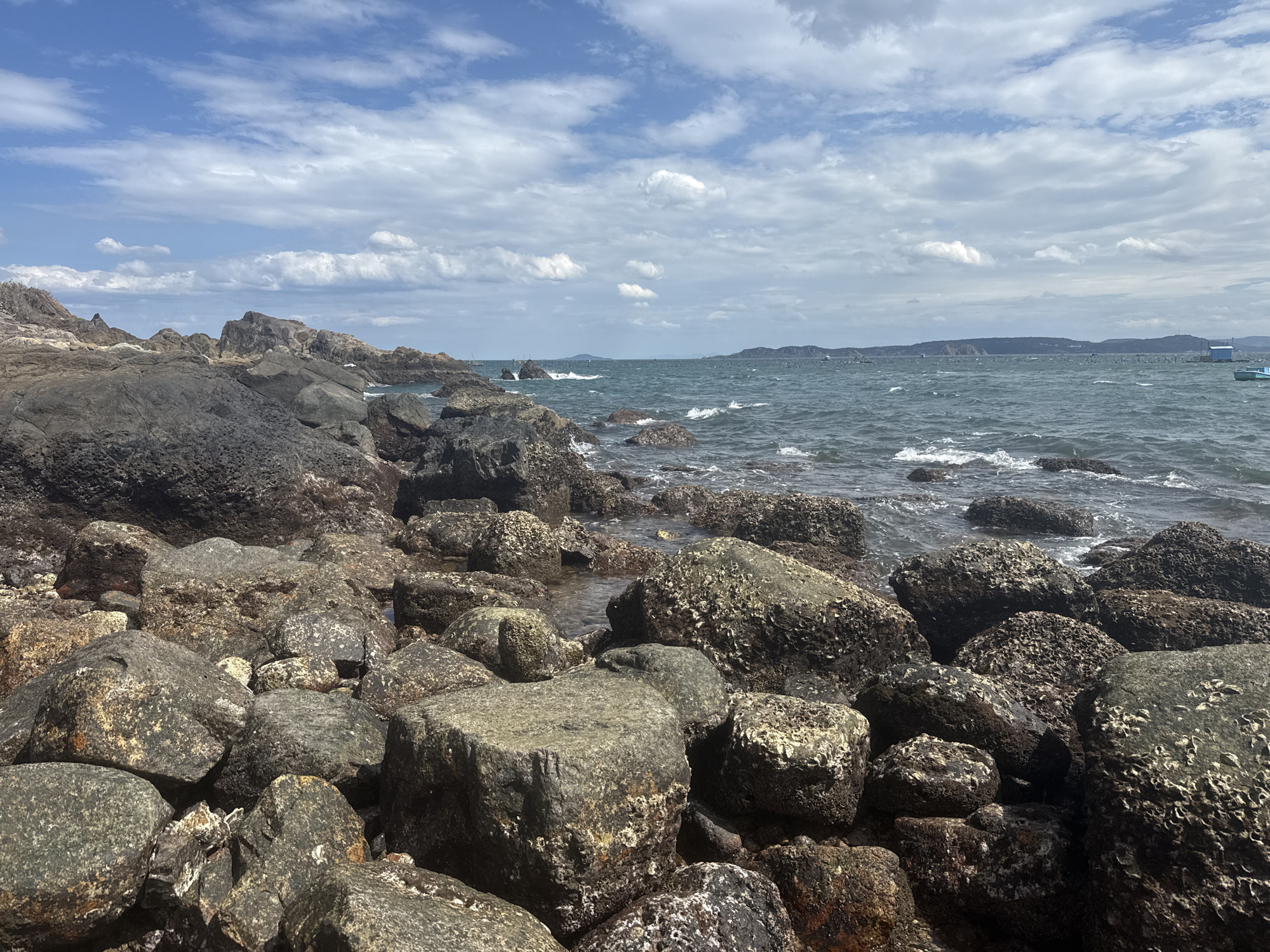 The image depicts a rocky coastline under a cloudy sky. In the foreground, a multitude of large, weathered rocks of varying sizes and textures cover the shore. Some rocks are dark and jagged, while others are lighter and rounded, with some showing signs of marine growth. The water is a deep blue-green and moderately choppy, with small waves breaking against the rocks. In the distance, a hazy shoreline with hills and some scattered structures can be seen. A few small boats are visible on the water. The overall impression is of a rugged and natural coastal landscape. This scene is characteristic of the beaches in Song Cau City, Phu Yen Province, Vietnam, which are known for their dramatic rocky formations and clear waters.
