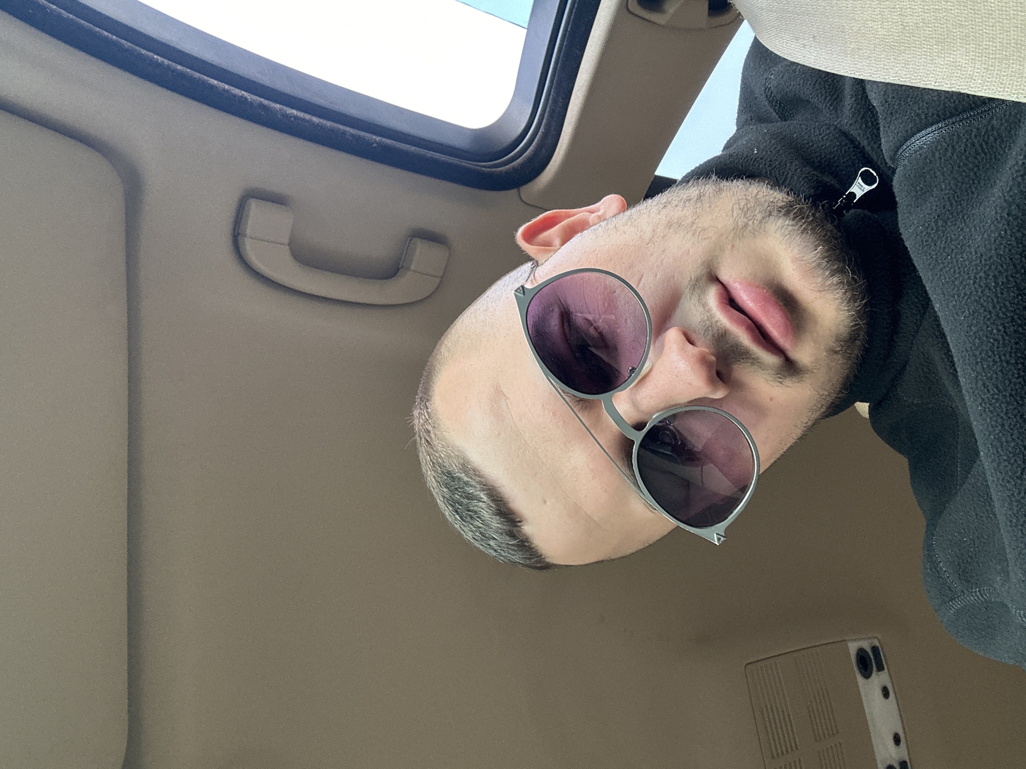 A man with sunglasses is in a car, presumably in Plovdiv, Bulgaria, given the prompt's instruction. The man has short, dark hair and is wearing a black fleece jacket. He is positioned close to the camera, which is angled upward, capturing a view of his face, a portion of his upper body, and the interior of the car. The car's interior features a sunroof, a passenger-side grab handle, and a beige interior. The light suggests it is daytime.