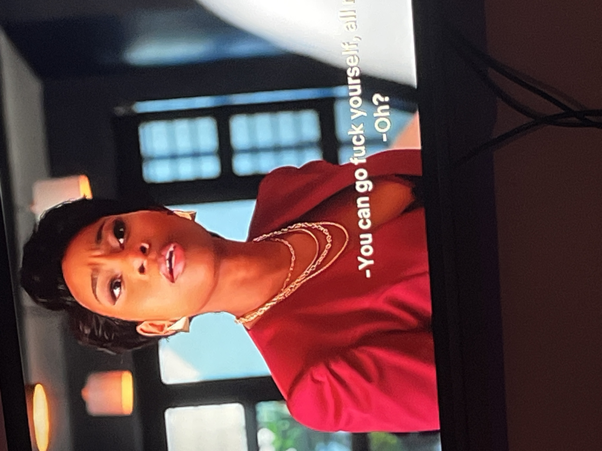 A close-up, rotated shot shows a Black woman with short hair, wearing a red top and gold necklaces. She has large, gold hoop earrings and appears to be speaking with her mouth open. The background is blurred, but suggests an indoor setting with what looks like windows with a grid pattern and interior lighting.
White text is superimposed on the right side of the image, oriented vertically. It reads: "-You can go fuck yourself, all r -Oh?". The text is partially obscured by shadows and the rotation of the image. The overall mood appears to be intense or confrontational, given the woman's expression and the explicit nature of the text. The Gura Top, Nigeria location context is not directly visible in the image itself.