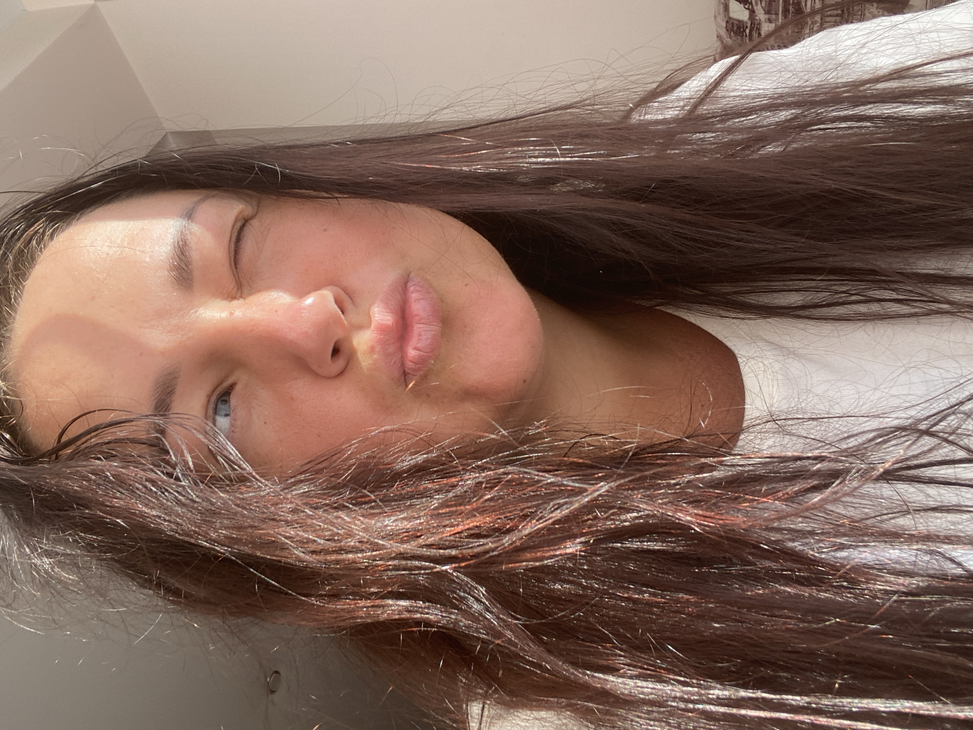 The image shows a close-up of a woman's face from the chest up. She has long, dark brown hair that appears slightly wet and is catching the light, giving it a reddish-brown sheen. Her hair is framing her face, with some strands falling across her forehead and cheek. She is wearing a white t-shirt.
Her facial expression is playful, with one eye closed in a wink and her lips pursed in a kissing gesture. Her skin is fair, and there appears to be some natural redness or flush on her cheeks. Sunlight is casting a diagonal shadow across the left side of her forehead and cheek, highlighting the texture of her skin and the strands of her hair. Her right eye, partially visible, is blue.
The background is out of focus, but it appears to be an indoor setting with neutral-colored walls and a window or bright light source to the right, suggesting daylight. There is a hint of a patterned object in the far right background, possibly furniture. The scene does not contain any specific indicators of the city of Sochi, Russia, or any other identifiable urban or natural landscape features.