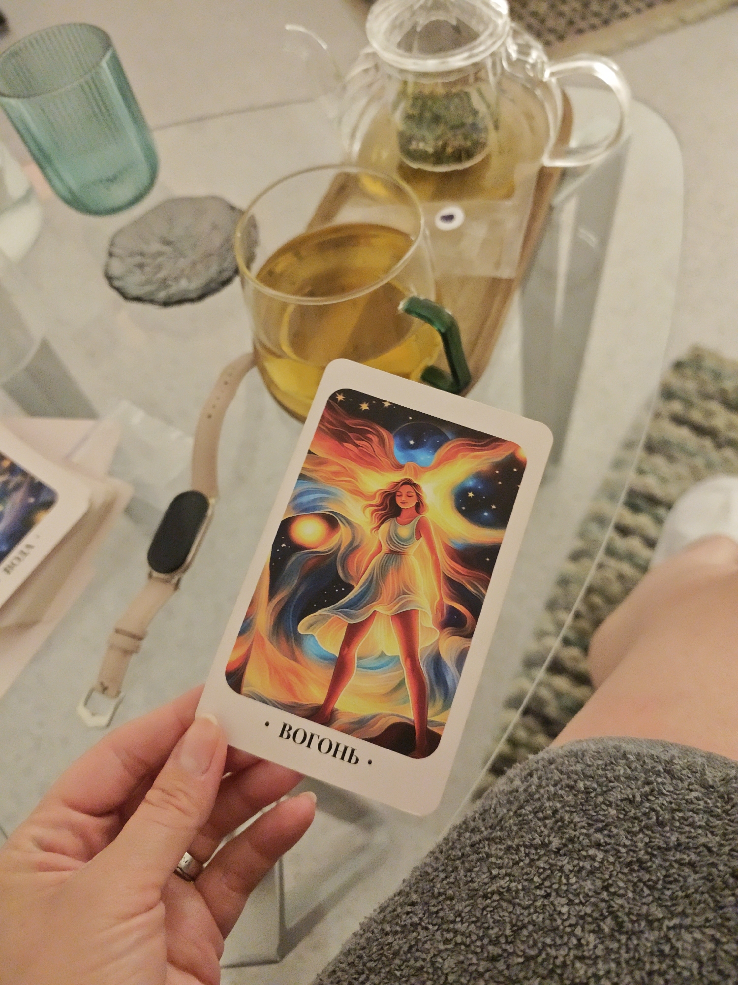 A hand holds a tarot card displaying a woman radiating light amidst celestial swirls. In the background, a glass teapot with tea and a teacup sit on a glass table, along with a smartwatch and other cards. The scene occurs on a soft, neutral-toned surface, possibly a rug or a couch. No specific details indicate the location is in Vinnytsia, Ukraine.