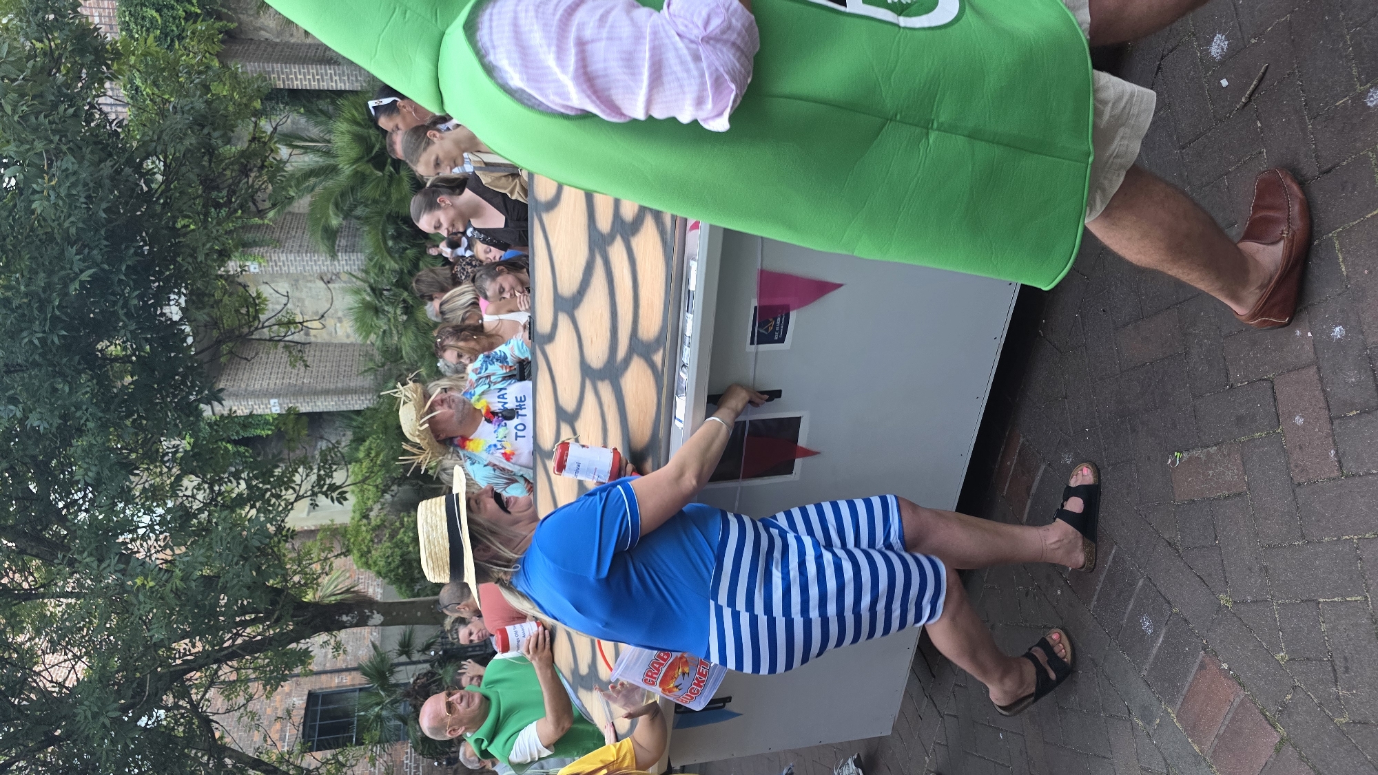 In St Leonards, United Kingdom, a woman wearing a blue and white striped dress, a straw hat, and a fake mustache is pushing a large, cardboard structure on wheels that is decorated to resemble a fish's scales. She is holding a red can. Behind her, a man in a green costume with a pink striped shirt is visible. A crowd of people in casual attire is gathered, some wearing Hawaiian shirts and leis. The scene appears to be an outdoor event or parade, with brick paving and trees in the background.