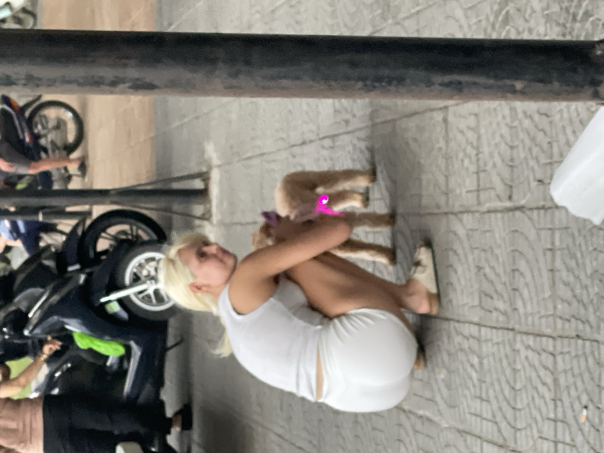 A young Caucasian woman with blonde hair is crouched down on a paved sidewalk, hugging a small, light-brown dog. The woman is wearing a white tank top and white shorts. The dog is wearing a bright pink collar. To the left of the woman, a dark-colored motorcycle is parked. Beyond the motorcycle, a bicycle is visible. The ground is paved with a light-colored, patterned tile. A dark metal railing runs horizontally across the upper portion of the image. The scene appears to be outdoors, likely in an urban setting. The lighting suggests it could be daytime, though the overall image quality is somewhat blurry and the orientation is rotated. There is no visible text in the image.