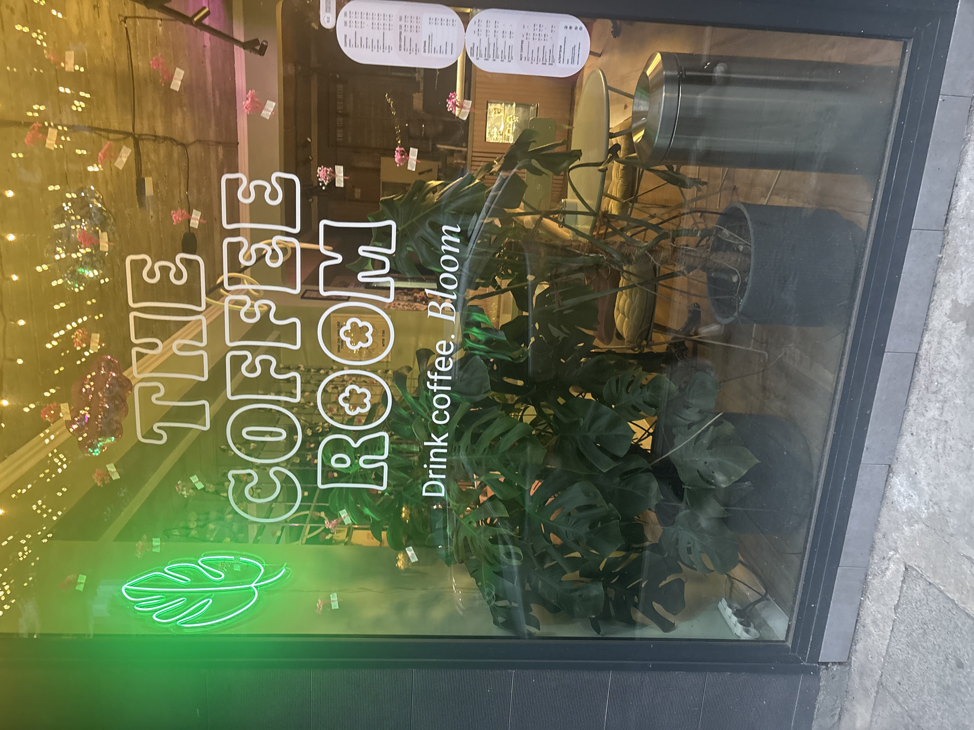 The image shows the window of a cafe called "THE COFFEE ROOM" in Burgas, Bulgaria. A neon green monstera leaf sign is visible on the left side of the window. Large white lettering on the window reads "THE COFFEE ROOM" and below that, "Drink coffee. Bloom." Inside the cafe, large potted plants, including a monstera, are visible. The interior appears to be decorated with string lights. A metal trash can and tables and chairs can also be seen through the window. Several menus are posted on the right side of the window. The exterior shows a grey tiled sidewalk.