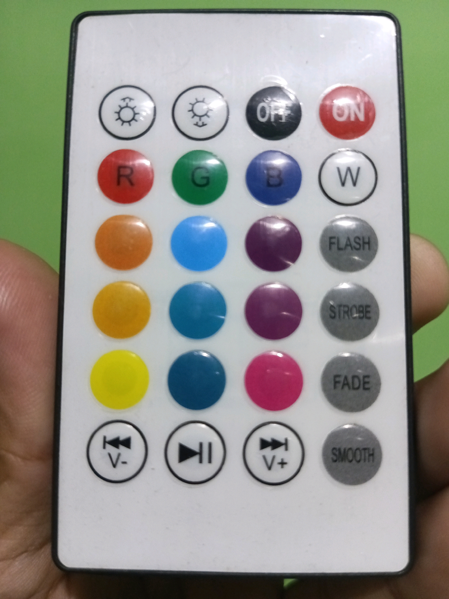 The image shows a close-up of a white remote control held in a person's hand against a plain green background. The remote has multiple colored buttons arranged in rows and columns, as well as some grey buttons with text labels. The colored buttons include red, green, blue, orange, yellow, purple, and pink, each with a letter or symbol on it. The grey buttons have labels like "OFF," "ON," "W," "FLASH," "STROBE," "FADE," and "SMOOTH." There are also buttons with play/pause and volume symbols. The remote appears to be for controlling LED lights or a similar electronic device. The lighting is even, and the focus is sharp on the remote.