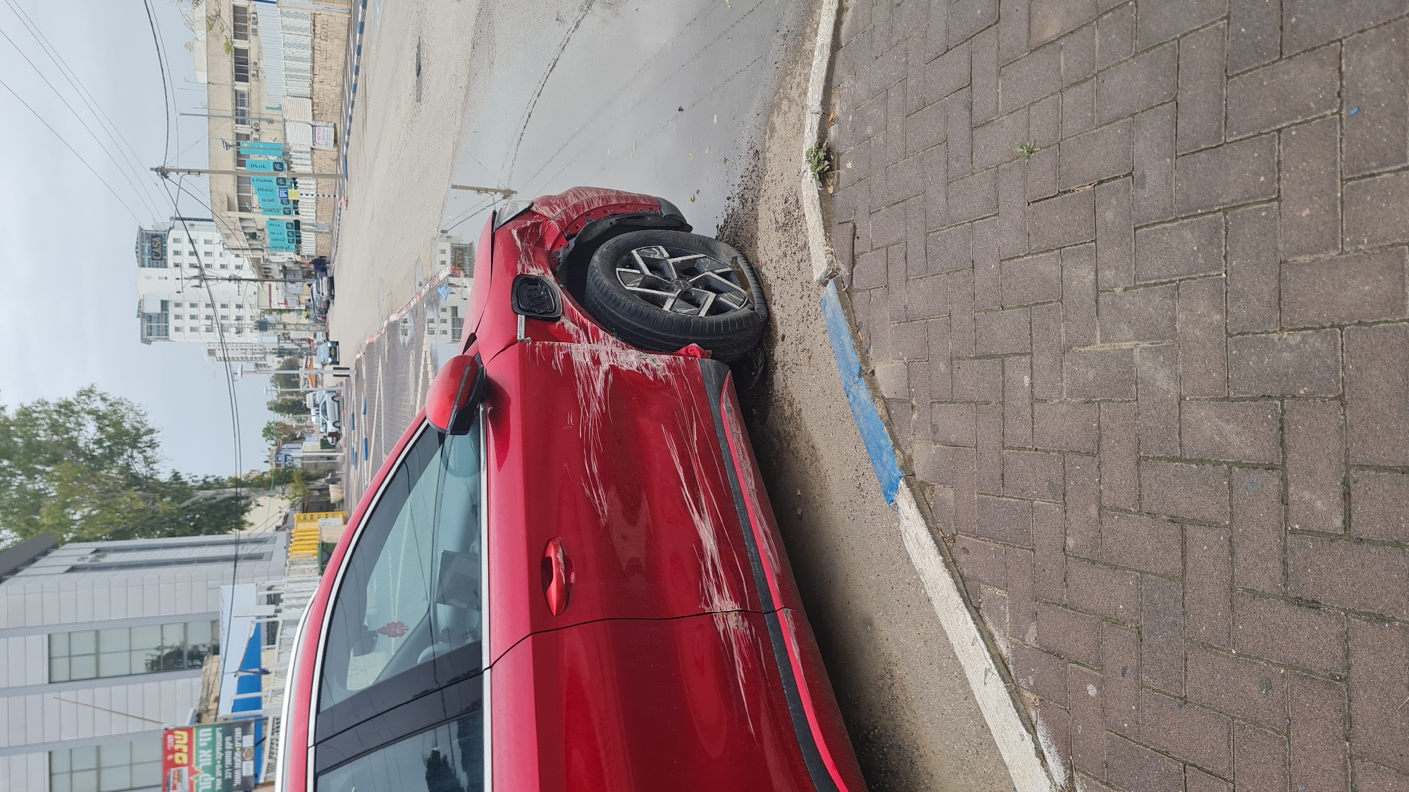 The image depicts a red vehicle that appears to have been involved in an accident. The car is positioned on the side of a street in Nahariyya, Israel, near a brick sidewalk with a blue painted curb. The vehicle's front driver-side tire is damaged and pushed inward. There are extensive scrapes and damage along the driver's side panels. The surrounding environment includes buildings, utility poles, and other vehicles, suggesting an urban setting. The sky is overcast. No individuals are immediately visible in the frame.