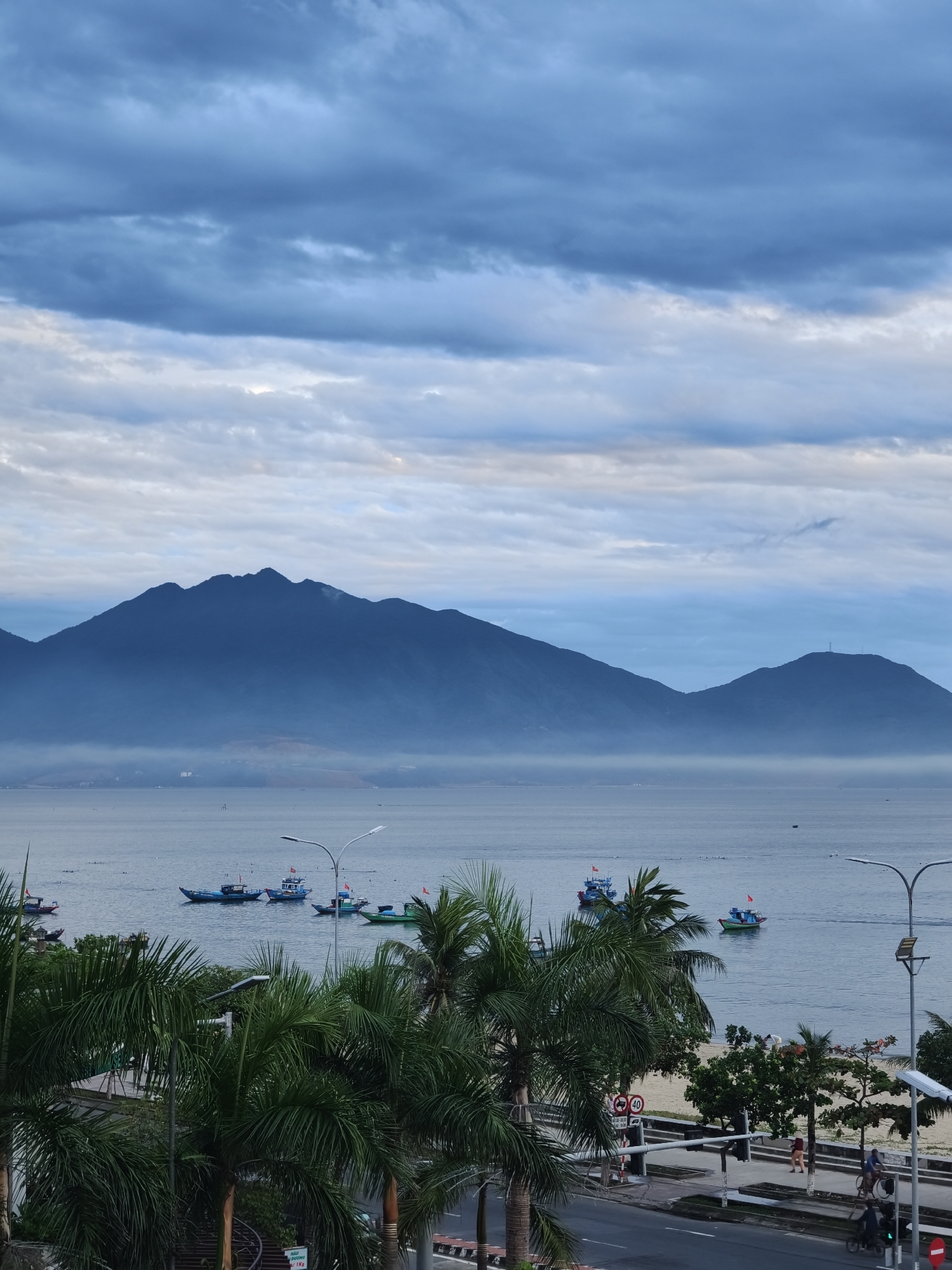 This is a photograph of the coastline in Đà Nẵng, Vietnam. In the foreground, palm trees line a street and sidewalk adjacent to a sandy beach. A road runs parallel to the sidewalk, with a few vehicles and pedestrians visible. Beyond the beach, the calm sea stretches out, dotted with several small fishing boats, some of which are adorned with red flags. In the distance, a range of blue, misty mountains dominates the horizon under a cloudy sky. The overall atmosphere is serene, suggesting an early morning or late evening scene.