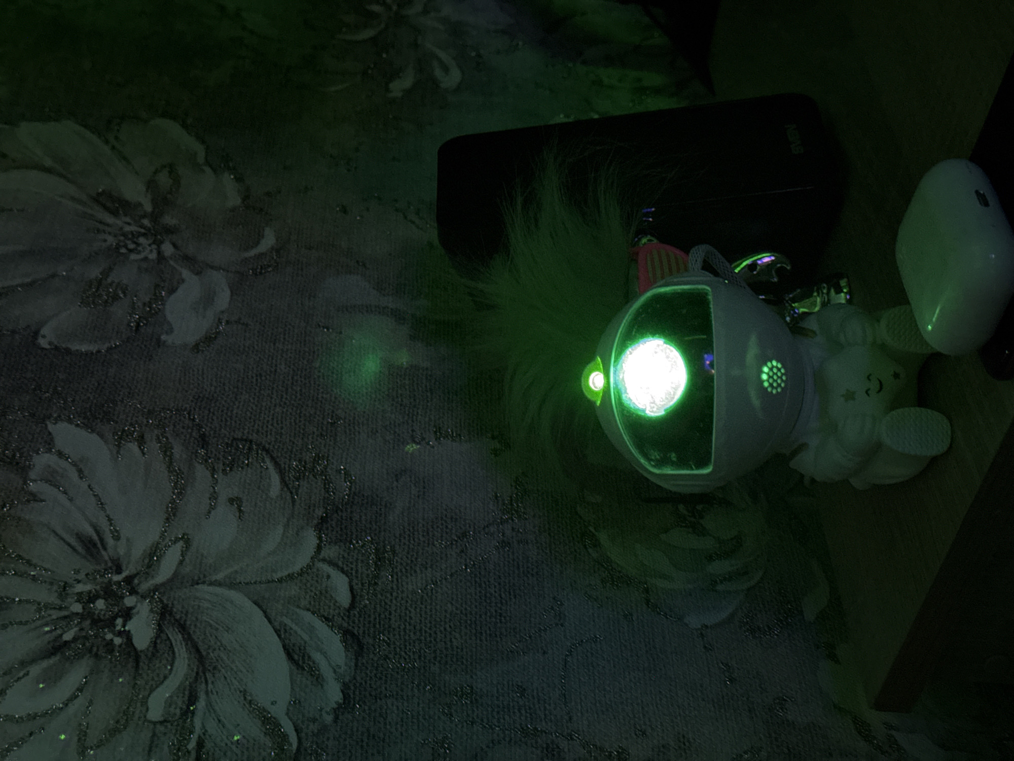 The image captures a dimly lit indoor scene, likely at night, with objects resting on a dark, patterned surface. The primary subject is a white, rounded electronic device emitting a bright green light from its central lens and a smaller green light to its left. It features a speaker-like grill and what appears to be green, fluffy material resembling hair or a pompom on its top.
To the right of this glowing device, a white, oval-shaped object, possibly a wireless earbud case, is visible. Further right, a small, white cartoonish figure, resembling a bear or similar animal, lies on its back, with a small star visible on its belly. Behind these items, a black rectangular object, possibly a phone or power bank, is positioned horizontally, with the brand name "SVEN" visible on its surface.
The objects are placed on a dark fabric surface adorned with a subtle, lighter floral pattern. Some areas of the pattern exhibit a textured or sparkly quality, catching the ambient light and the green glow from the device. The strong green light from the electronic device casts a distinct hue onto its immediate surroundings, creating illuminated spots and reflections on the dark background. No people are visible, and the scene depicts stationary objects.