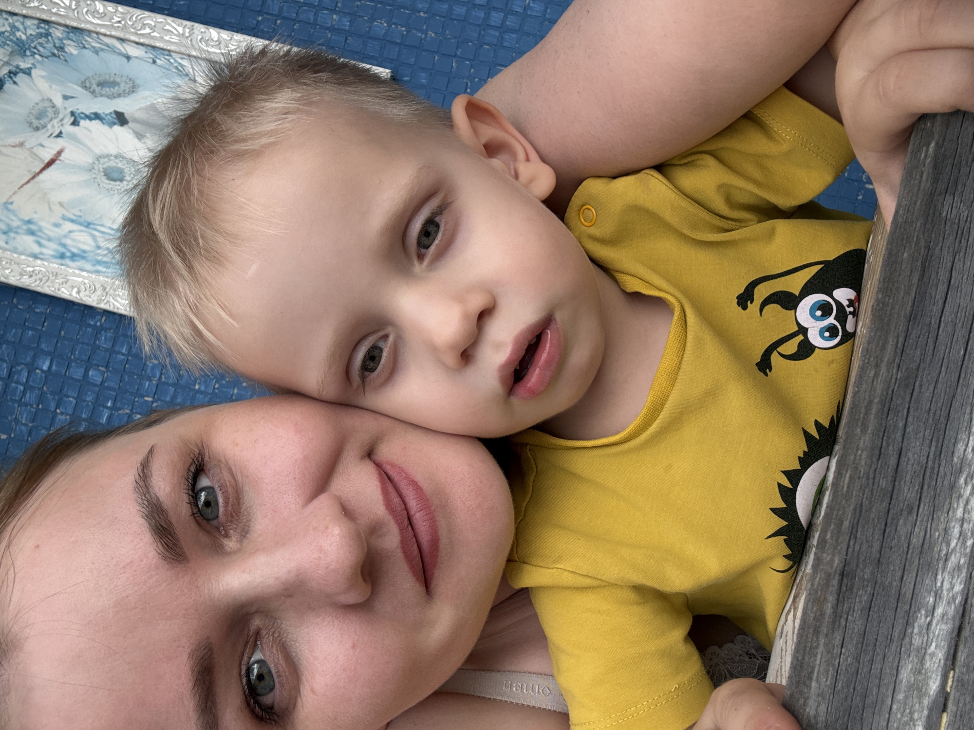 A close-up, head-and-shoulders outdoor portrait shows a woman embracing a young child, both looking toward the camera. The woman, with light brown hair pulled back and fair skin, has green eyes and is wearing a light-colored strap, possibly from a tank top. The child, with blonde hair and fair skin, has blue eyes and is wearing a bright yellow t-shirt with a cartoon monster graphic. The background consists of a textured blue surface, possibly siding or a wall, with a framed picture containing white and blue floral imagery partially visible. The lower portion of the image shows a wooden surface, likely a table or railing, in a gray, weathered tone. The image has been taken in the city of Chaikovsky, Russia.