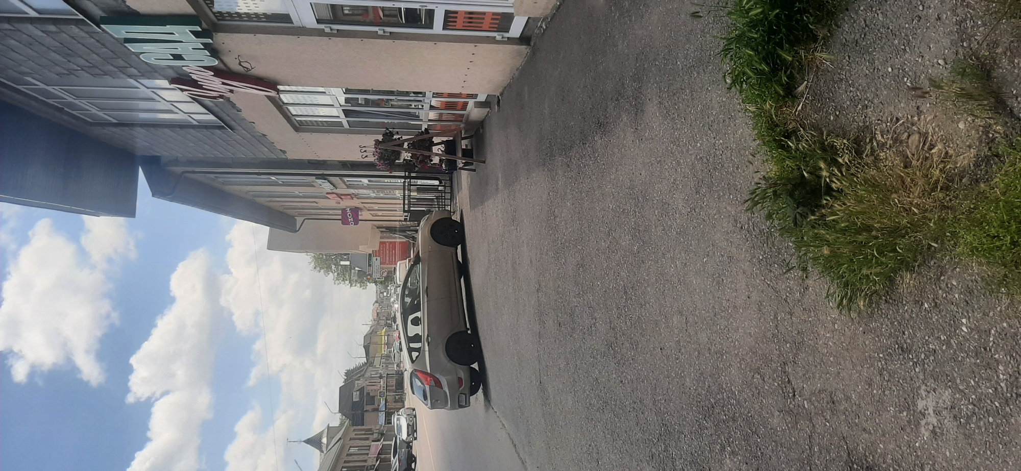 The image shows a street scene in Yessentukskaya, Russia. A beige hatchback car is parked on the side of the road, facing left. The car is a Hyundai Accent or similar model, with black rims. To the right of the car is a building with a light-colored facade and a dark grey mansard roof. The building has large windows, some of which are boarded up or have security grates. A sign above the windows reads "Yugo SAD" in green lettering. In front of the building, there is a wooden tiered stand with potted plants. A black metal fence is visible in front of the building. Further down the street, more buildings and cars are visible, suggesting a commercial or residential area. The sky is blue with scattered clouds. The ground is paved with asphalt and gravel, with patches of grass and weeds growing along the edge of the road. The overall impression is a casual daytime street view.