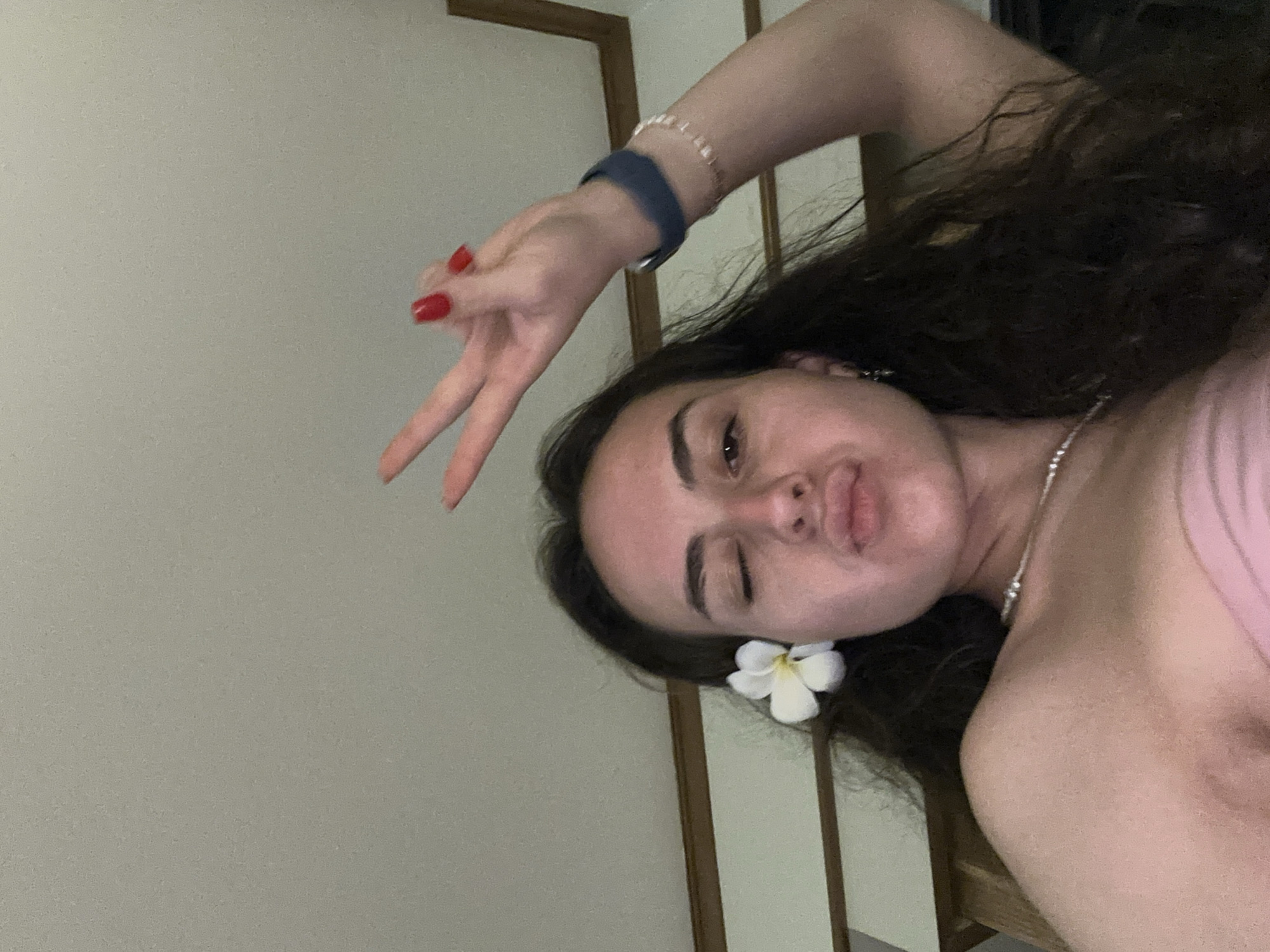 A selfie shot of a young woman, who is oriented horizontally and fills the majority of the frame. She has long, dark curly hair. She has a white flower tucked behind her ear. She is wearing a thin necklace. Her nails are painted red. She is making a peace sign with her fingers. She is wearing a dark blue watch on one wrist, and a string bracelet on the other. The background consists of a light-colored wall on the left, and a dark frame or doorway on the right. The environment is indoors. The location is Pattaya City, Thailand.