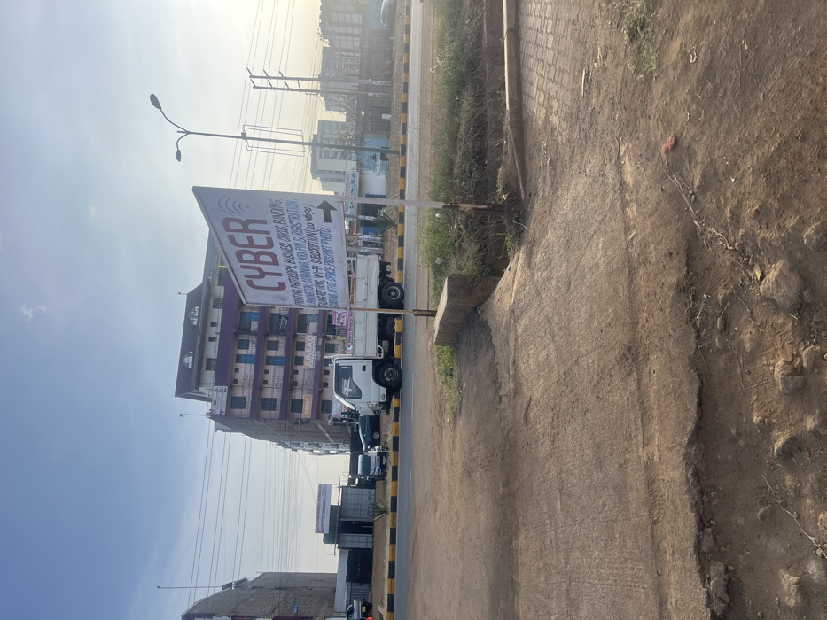 This media file captures a bright, sunny street scene in Gitothua ward, Kenya, likely during the morning or late afternoon. The main subject is a prominent directional sign for a "CYBER" cafe, made of white material with red text, featuring an arrow pointing to the right. The sign is positioned at the edge of a dirt and gravel path that runs alongside a paved road.
The services listed on the sign are:
* CYBER
* PRINTING
* PHOTOCOPY
* BUSINESS CARD BINDING
* COMPUTER TRAINING
* KRA PIN & REGISTRATION
* FILING RETURNS
* WI-FI SUBSCRIPTION (20 Mbps)
* RENTING OFFICE SPACE
* PASSPORT PHOTO
In the background, multiple buildings of varying heights and colors are visible, including a multi-story structure with purple and beige facades. Utility poles with numerous overhead wires extend across the sky. Several vehicles, including a white truck and other cars, are parked or driving on the paved road, indicating active traffic or commerce. The immediate foreground shows a rough, unpaved area with some scattered debris and sparse vegetation. The clear sky suggests fair weather.