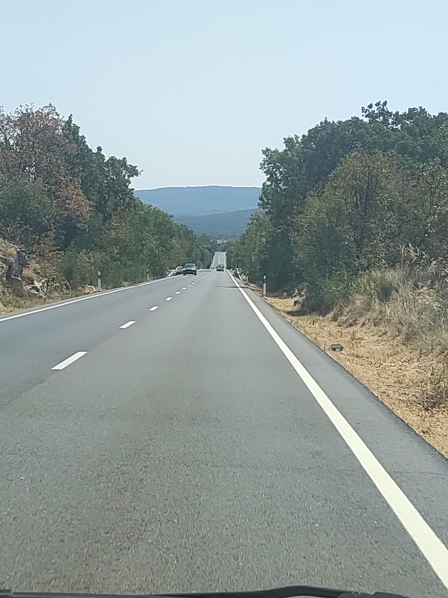 The image shows a two-lane asphalt road stretching into the distance, flanked by trees and dry vegetation. The sky is clear and pale blue. In the foreground, the road has a white solid line on the right edge and dashed white lines dividing the lanes. Several cars are visible on the road, heading away from the viewer. The landscape appears to be hilly, with distant mountains visible on the horizon. This scene is consistent with the roads found in the vicinity of Becerril de la Sierra, Spain, a municipality in the Community of Madrid known for its natural landscapes and proximity to mountainous regions like the Sierra de Guadarrama.
