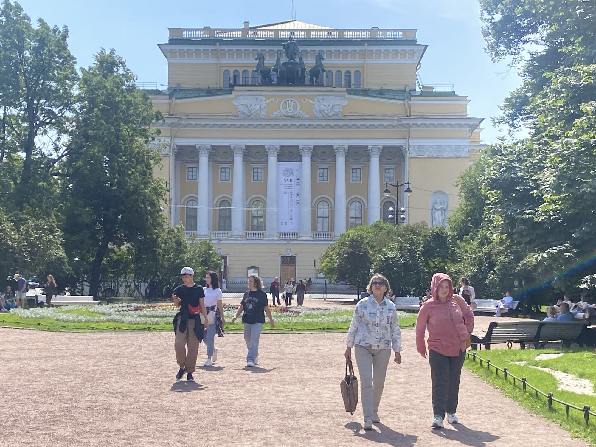 The image depicts a view of the Alexandrinsky Theatre in Saint Petersburg, Russia, on a sunny day. Several people are walking on a gravel path in the foreground, with a landscaped garden and the grand yellow building of the theatre in the background. The theatre features classical architecture with large columns, a prominent statue on its roof, and a banner hanging between two columns. Lush green trees frame the scene, and benches are visible in the park area.