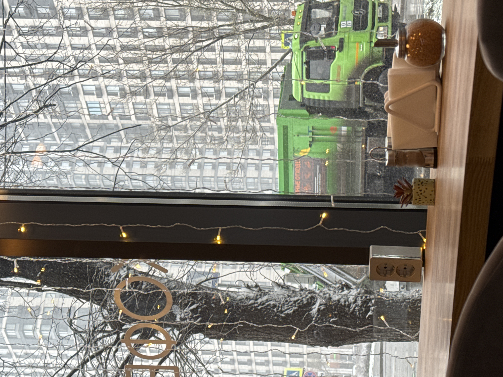 The image depicts a view from inside a building, possibly a cafe or restaurant, looking out onto a snowy street scene in an urban area. The focus is on a window frame decorated with string lights, which frame the external view of a large building and trees covered in snow. A green truck is visible on the street outside.
On the right side of the frame, there is a table with a napkin holder and a sugar dispenser. A power outlet is visible mounted near the window frame. The overall impression is of a cold, snowy day outside, contrasting with the warm and cozy interior.