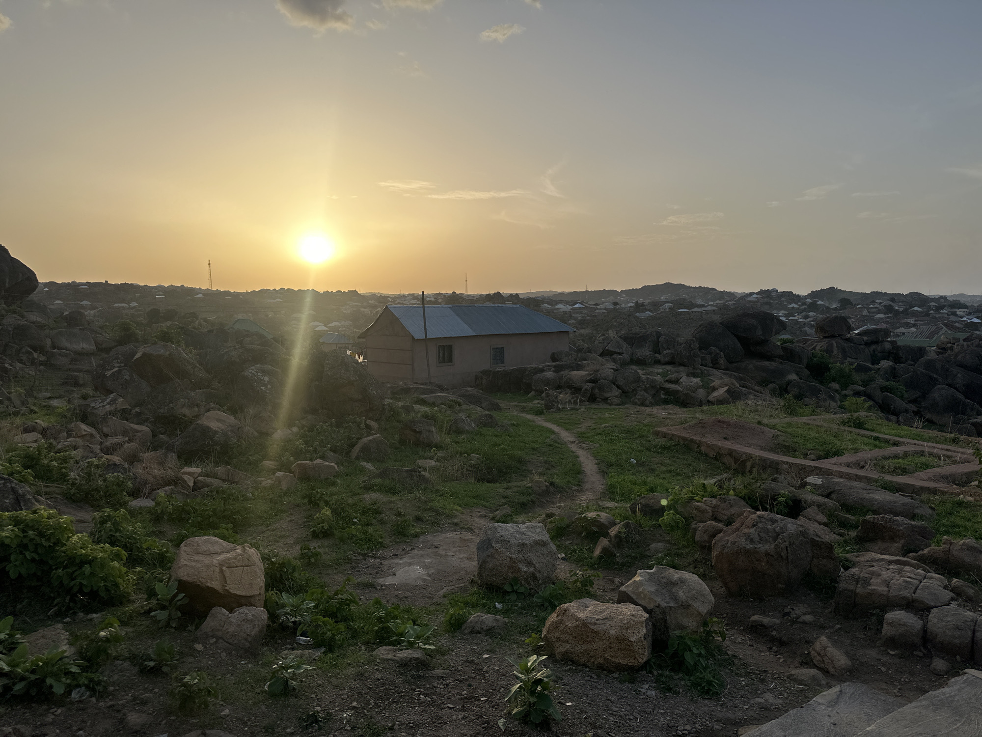 The image depicts a landscape in Jos, Nigeria, featuring a building nestled among rocky terrain. The sun is setting, casting a golden light across the scene. The foreground is dominated by rocks and sparse vegetation, with a dirt path leading towards the building. The building has a metal roof and small windows. In the background, the city of Jos is visible as a cluster of buildings against the horizon. A communication tower can be distinguished among the city skyline. The overall atmosphere is peaceful and rural, despite the proximity to the city.