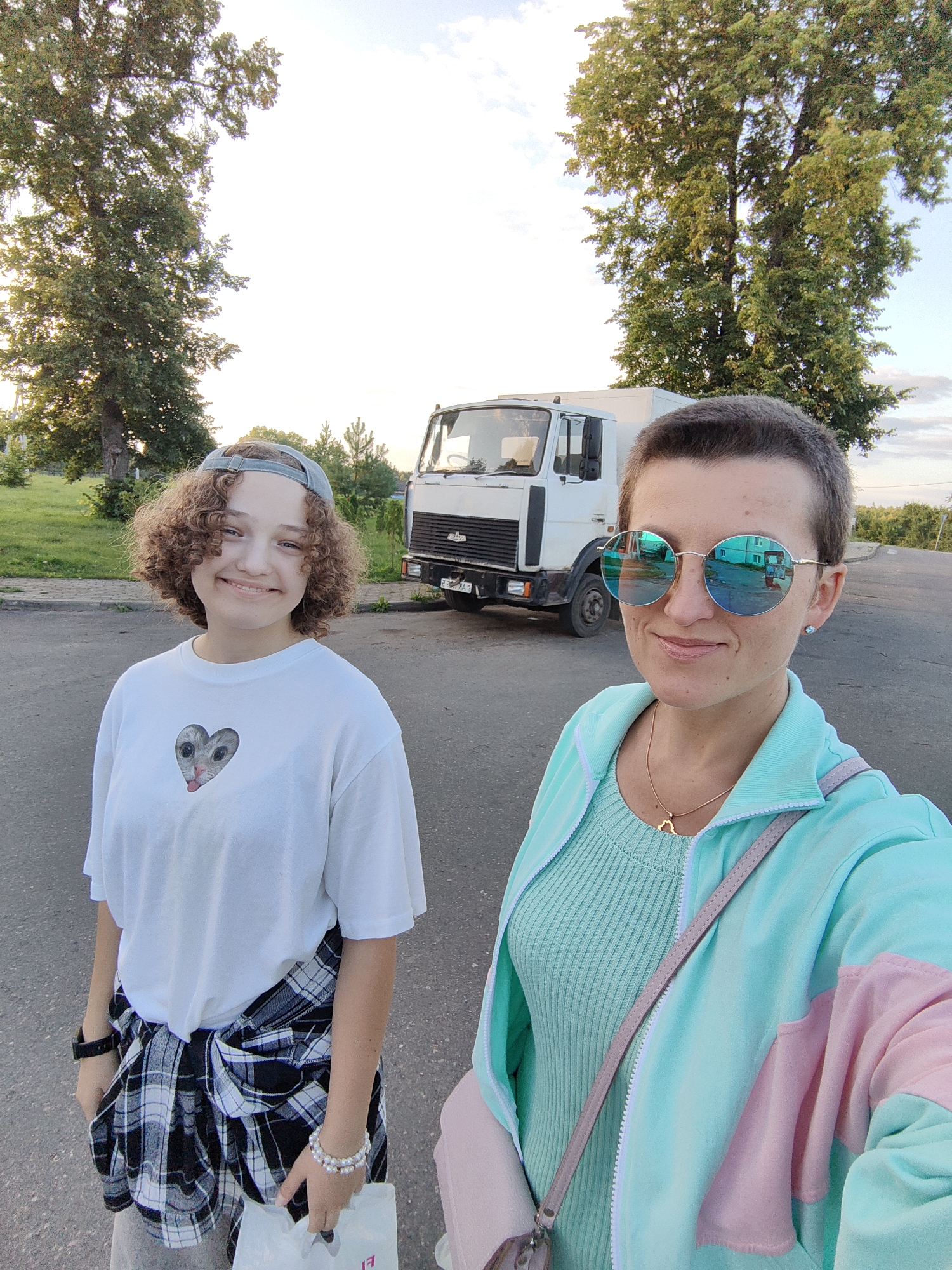 Two individuals stand in an outdoor urban setting, with a white truck in the background. The person on the left is a young female with curly brown hair, wearing a white t-shirt with a heart-shaped cat graphic, a grey baseball cap, and a plaid shirt tied around her waist. She is holding a white paper bag. The person on the right is an adult female with short dark hair, wearing large round sunglasses with blue mirrored lenses, a mint green ribbed top, and a teal and pink color-blocked jacket. She has a necklace with a star pendant and is carrying a light pink bag. The environment includes a paved road, grass, and large trees. The sky is visible, suggesting daytime with possibly late afternoon lighting. The license plate of the truck appears to be Belarusian. The location is Myadzyel, Belarus.
