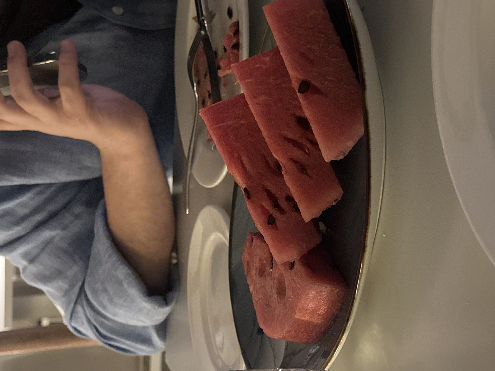 The image shows a close-up view of sliced watermelon on a dark plate. The watermelon slices are triangular and have seeds visible. In the background, a person wearing a blue shirt is visible, with their arm and hand holding a cell phone. The setting appears to be a restaurant or dining area, with plates and cutlery visible on a table. The lighting suggests an indoor environment, possibly a dimly lit room. There are no specific visual cues in the image that would indicate the location is Yerevan, Armenia.