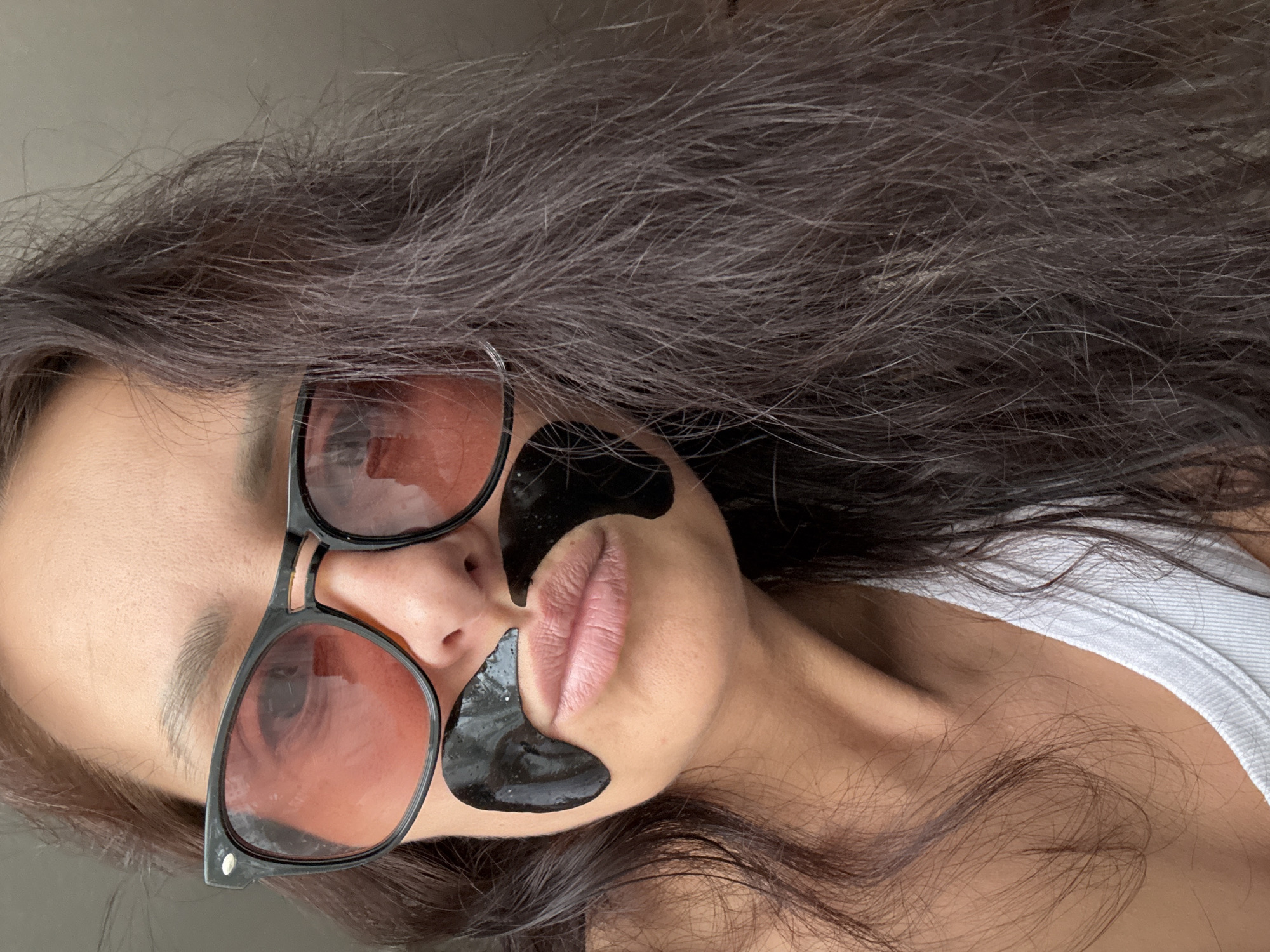 A woman with brown hair and light skin is lying down, facing the camera and rotated counter-clockwise. She is wearing black-framed sunglasses with rose-tinted lenses, as well as black under-eye gel patches. Part of a white tank top is visible near her shoulder. The location is not discernible from the image.