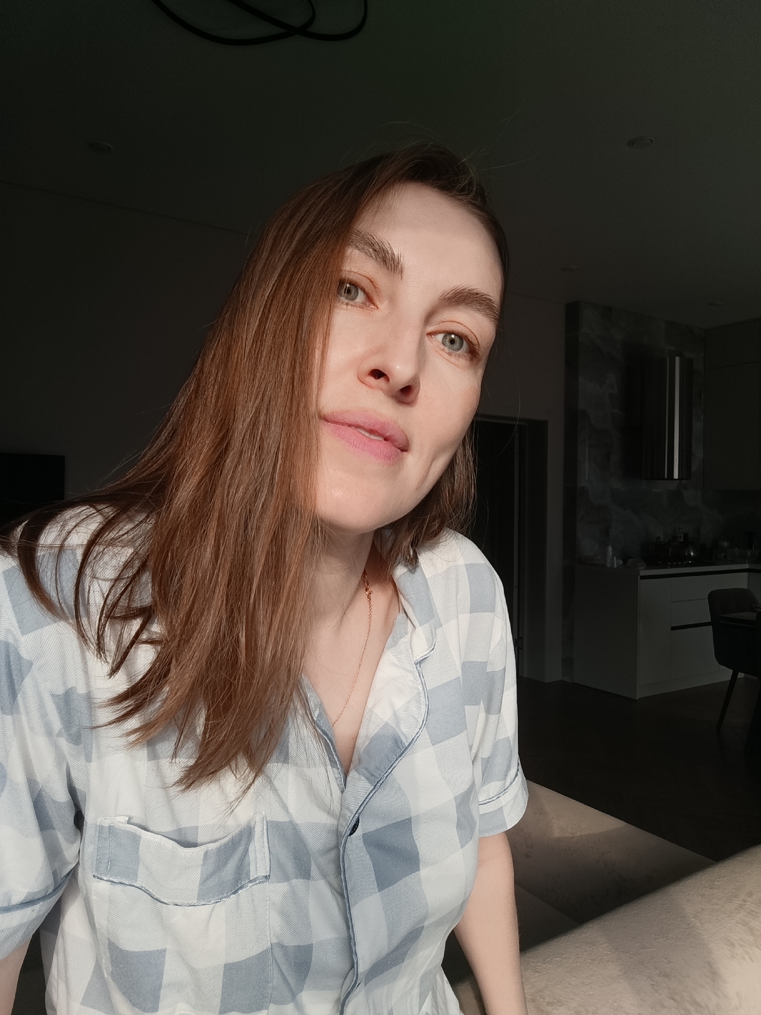 A young woman is indoors, likely in an apartment. She has light skin, brown hair, and light-colored eyes. She is wearing a blue and white checkered shirt. The background shows a partially visible kitchen area with dark cabinetry and countertops. The lighting suggests late afternoon or early morning with a strong directional light source. The location is Chaikovsky, Russia.