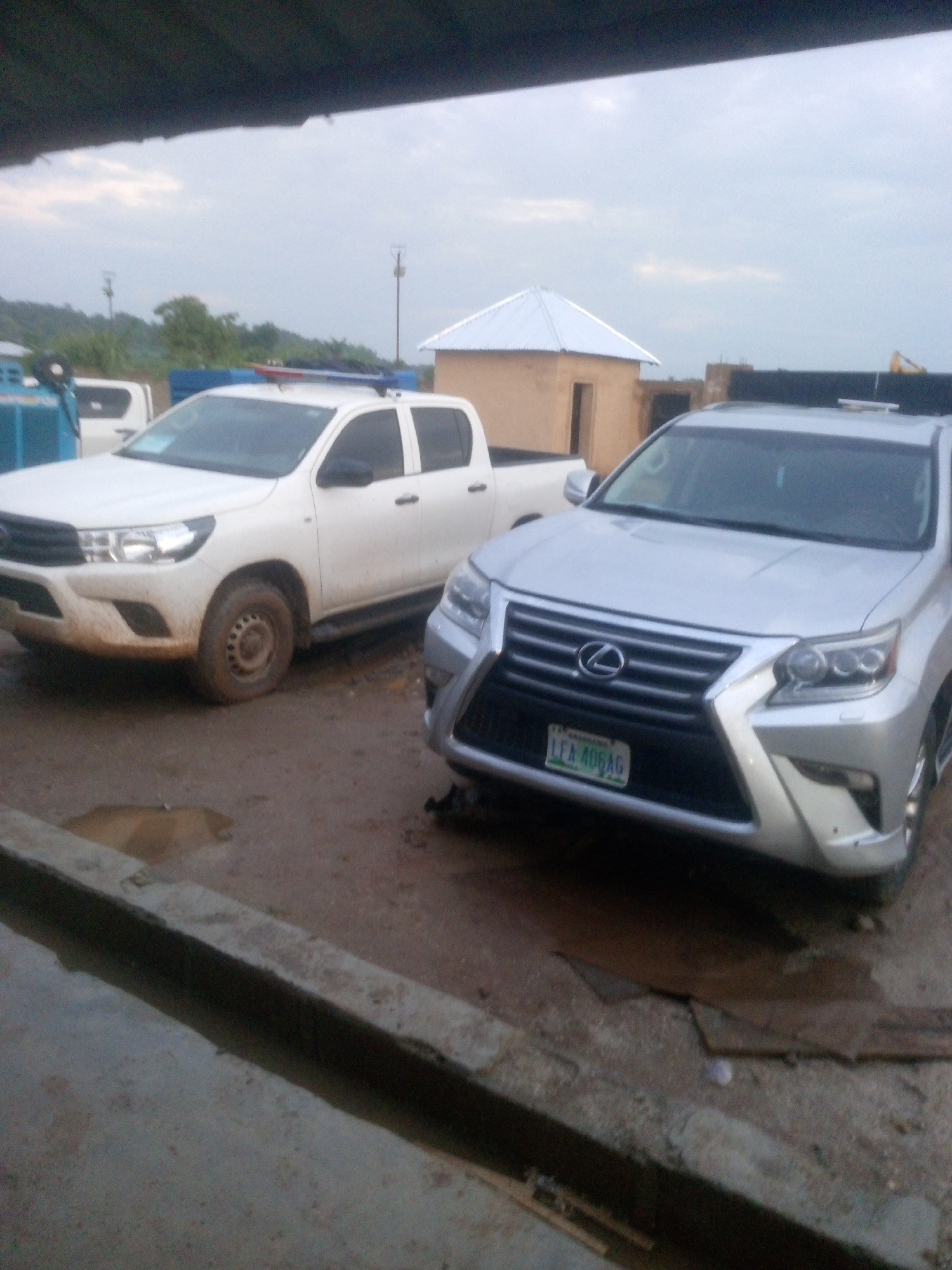 The image depicts an outdoor scene, likely in Indo, Nigeria, featuring two vehicles parked on a dirt and muddy ground. A white Toyota Hilux pickup truck is on the left, appearing to be a police vehicle with a red and blue light bar mounted on its roof. The truck is heavily soiled with mud. To the right is a silver Lexus SUV, also appearing to be a civilian vehicle, with a license plate that reads "LEA 406 AG". The Lexus is positioned in front of and slightly to the right of the Hilux.
In the background, a small, unfinished building with a corrugated metal roof is visible. Beyond that, there are trees and a cloudy sky. The ground is uneven and covered in mud, with puddles of water present, suggesting recent rainfall or a poorly drained area. The overall impression is one of a functional, possibly semi-rural or developing area, where vehicles are exposed to the elements and the ground conditions are challenging. No people are visible in the image.