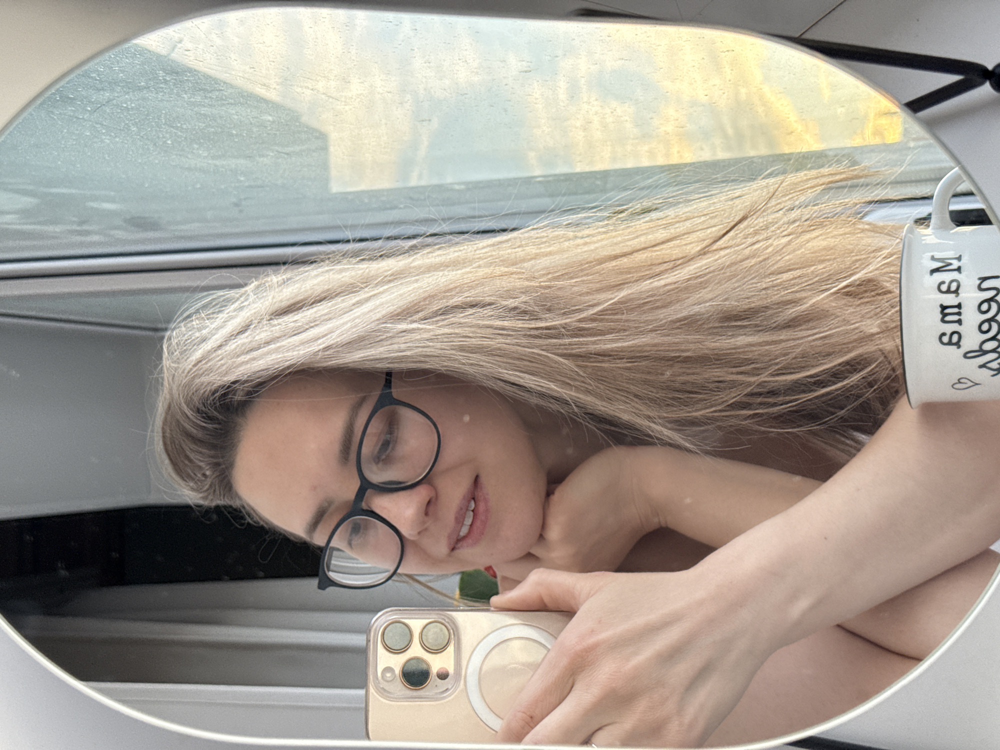 A mirror selfie of a blonde woman with glasses, holding a gold iPhone. The reflection shows a window with daylight visible, suggestive of early morning or late afternoon. The photo also includes part of a mug. The exact location in Chaikovsky, Russia cannot be determined from the image alone.