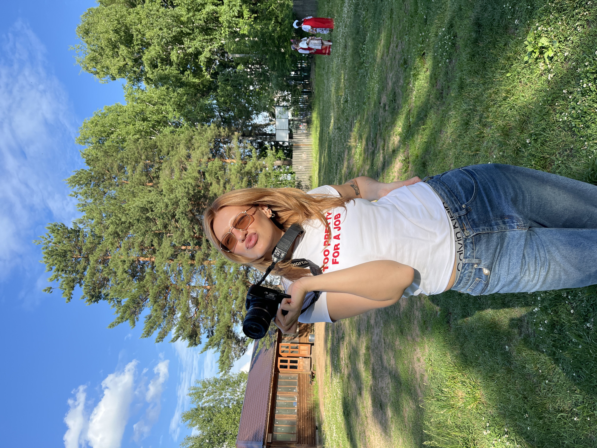 In a sun-drenched outdoor setting, a young woman with blonde hair and sunglasses poses for a selfie. She's wearing a white t-shirt with red text that reads "TOO PRETTY FOR A JOB" and blue jeans. A black digital camera is held in her hands, and a strap is around her neck.
In the background, two individuals in traditional red and white costumes are visible on a grassy field. A wooden building with a brown roof and windows stands to the left, partially obscured by trees. The scene is set against a backdrop of lush green trees and a bright blue sky with scattered clouds, suggesting it's daytime on a pleasant day. The overall mood appears casual and relaxed, with the woman looking directly at the camera with a playful expression. The location cues suggest Chaikovsky, Russia.