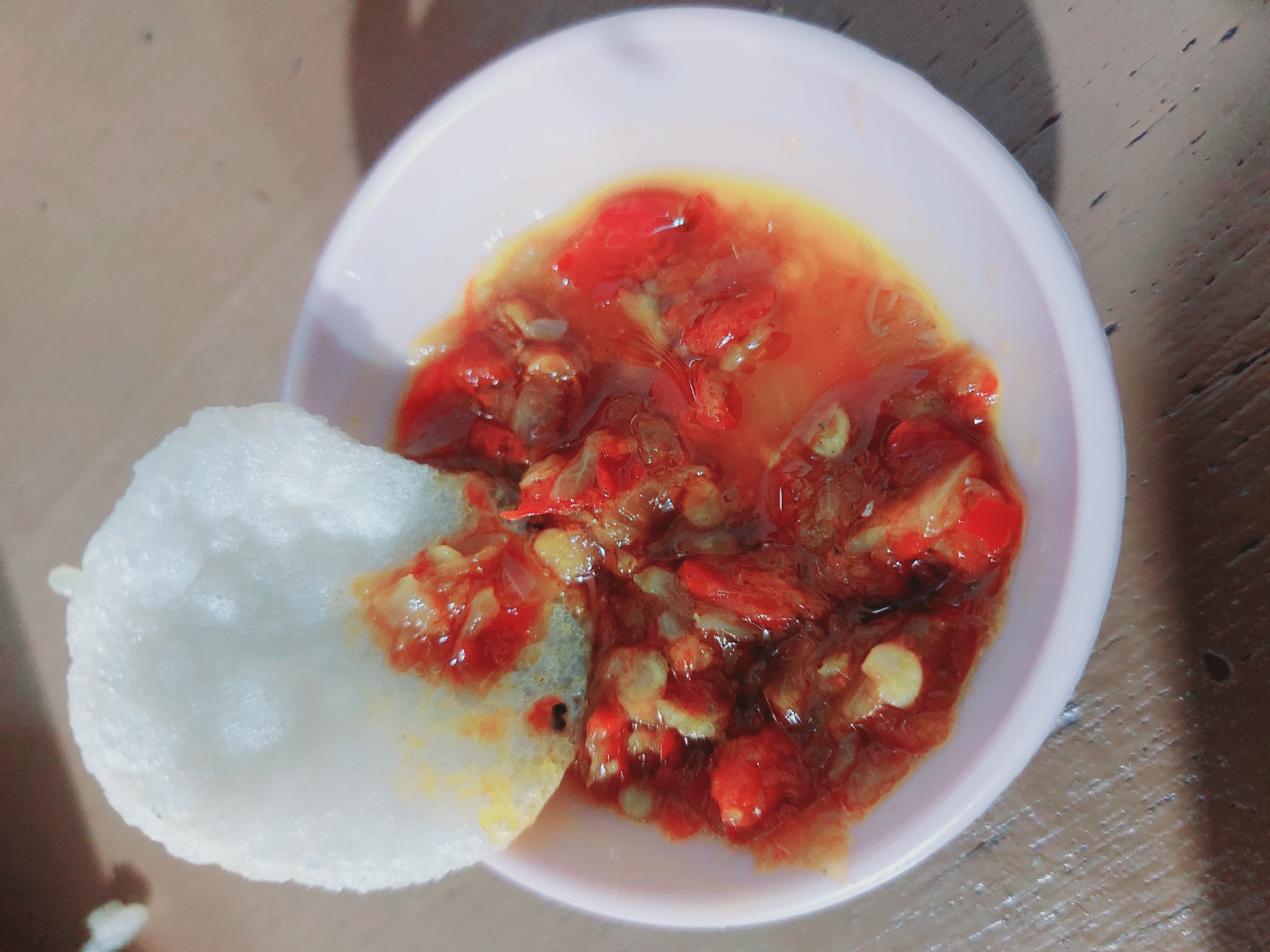 A close-up, overhead shot shows a small, light pink bowl filled with a vibrant red, chopped chili condiment. A large, white cracker with a bubbly texture is partially submerged in the condiment. The cracker's underside is coated in the red mixture, suggesting it has been dipped. The condiment appears to be made of finely chopped red chilies, possibly mixed with onions or garlic, and has a slightly oily sheen. The bowl rests on a wooden surface with a visible grain pattern. The overall lighting is soft, creating some shadows and highlighting the glossy texture of the condiment. The scene is simple and focused on the food item, with no people or other objects clearly visible within the frame. The image does not contain any specific visual cues to determine its location in Nunukan, Indonesia.