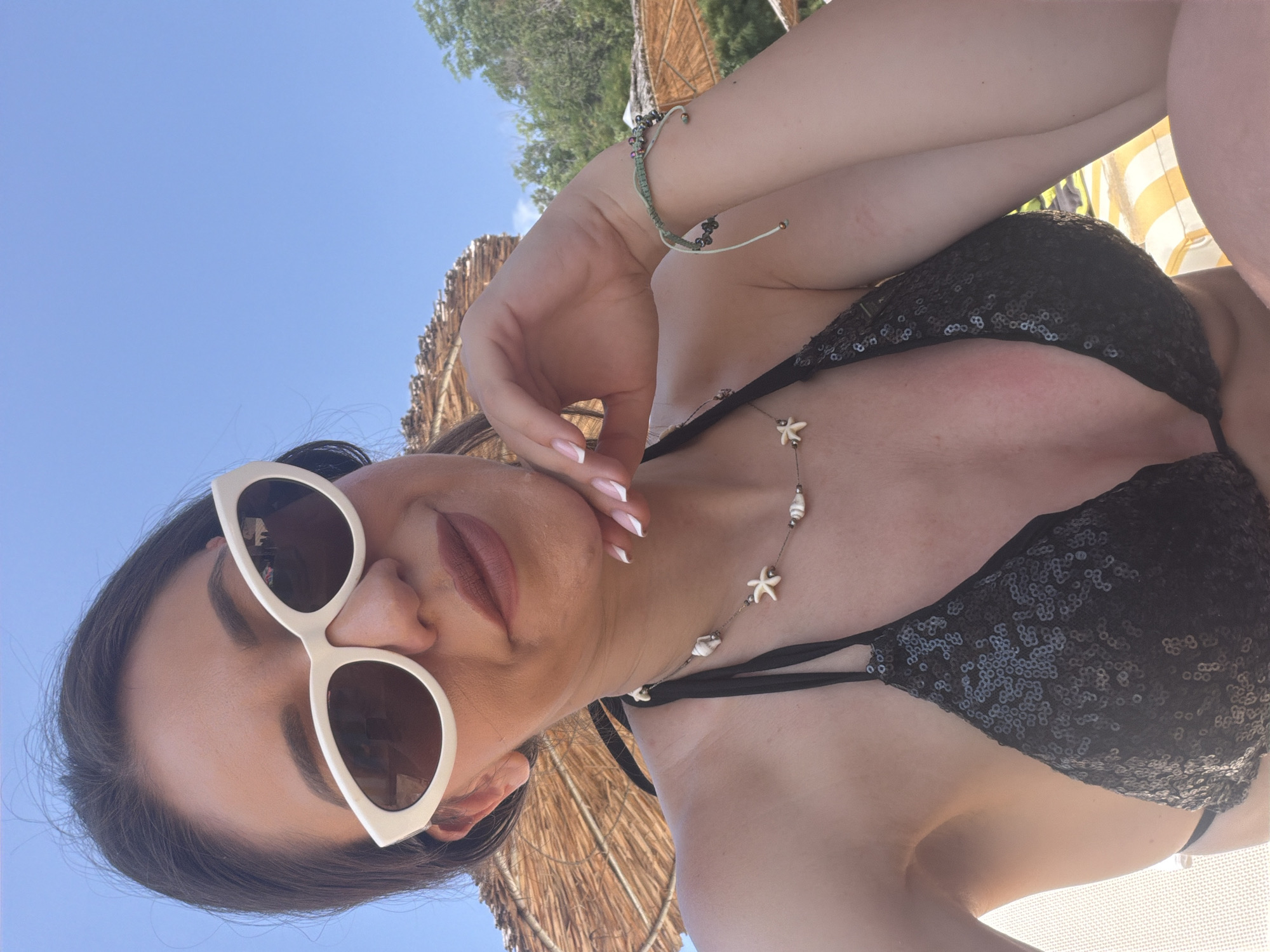 A woman with dark hair, wearing white cat-eye sunglasses, a black sequined bikini top, and a seashell necklace, poses for a selfie outdoors. She has her hand on her chin and her nails are painted with a French manicure. A green bracelet with beads is on her wrist. In the background, there are thatched umbrellas and trees. The sky is blue. The setting appears to be a beach or resort in Varna, Bulgaria.