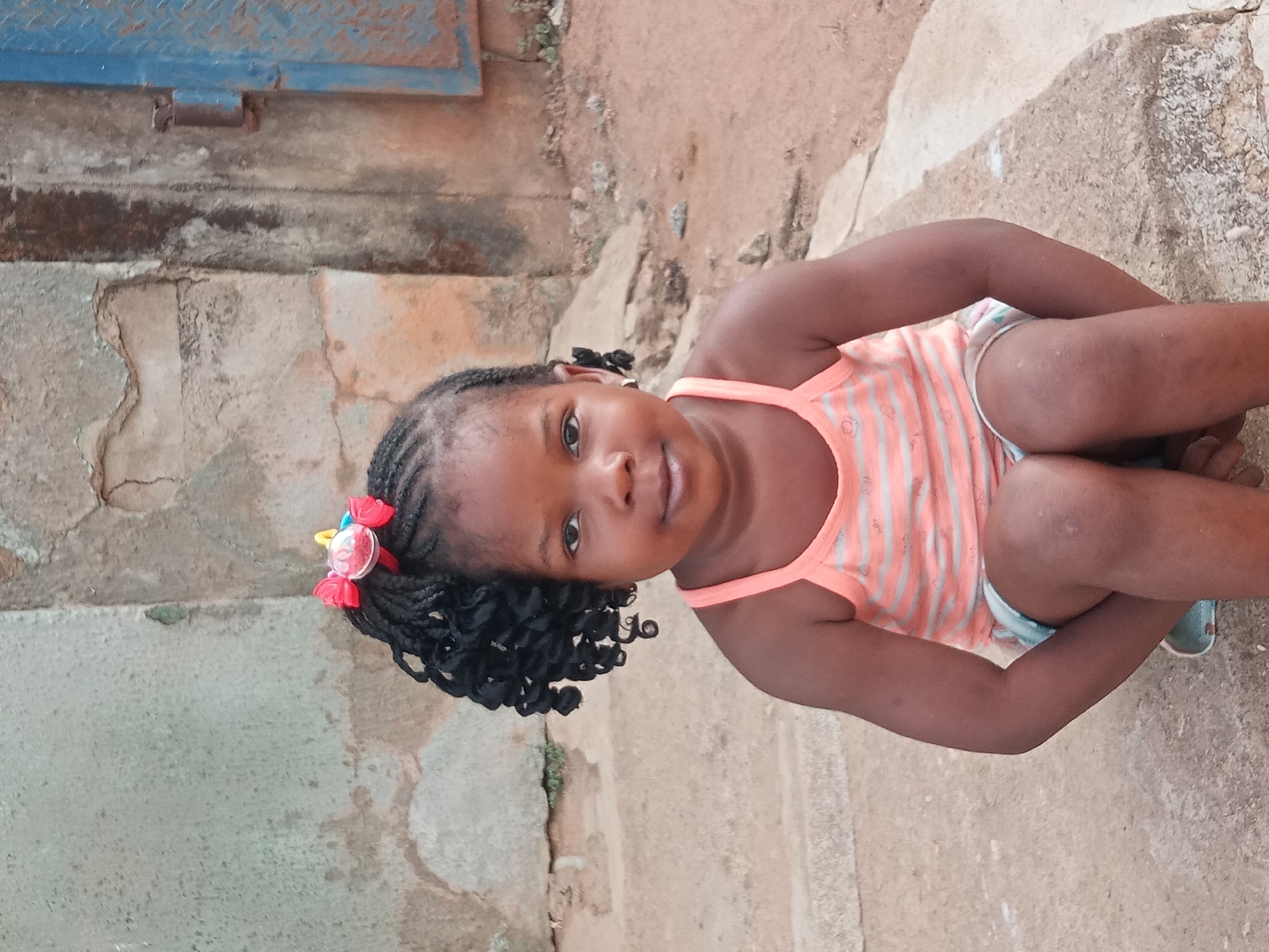 A young girl is sitting on a weathered, concrete surface. She has dark skin, braided hair with curled ends, and a decorative hair clip with red, yellow, and pink elements. She's wearing a light orange and white striped tank top. The background shows a worn, textured concrete wall or surface to her left, possibly part of a building. To her right, the concrete surface blends into a dirt or sandy area. The image is taken at an angle, suggesting the photographer was standing or kneeling. This is Sabon Tasha, Nigeria.