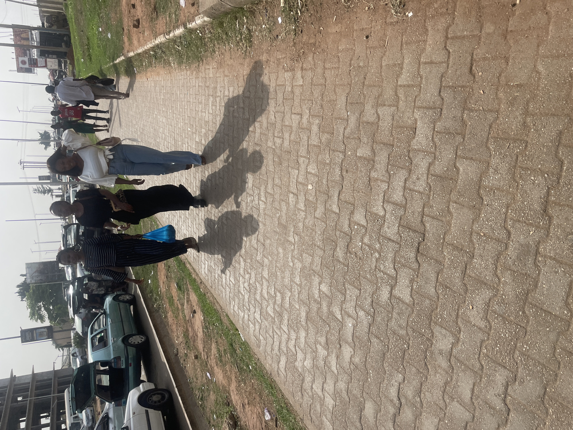 This daylight image captures a bustling street scene along a paved sidewalk in Abuja, Nigeria. The photograph is taken from a slightly elevated perspective, looking down the length of the sidewalk.
Several pedestrians are walking on the interlocking block pavement. In the mid-ground, a woman wearing a white long-sleeved top and blue jeans walks towards the left, carrying a light-colored shoulder bag. Her elongated shadow, along with others, is cast prominently on the sidewalk, indicating that the sun is low in the sky, suggesting either morning or late afternoon. Closer to the foreground, a woman dressed in a black top or dress walks, and just beyond her, another woman in a black and white striped outfit carries a blue bag. Other individuals are visible further down the sidewalk, some less distinct.
Adjacent to the sidewalk is an unpaved strip of dirt and sparse green grass, beyond which a busy road with numerous parked cars is visible. A light blue or green car with its boot open is noticeable among the vehicles. In the background, utility poles, distant buildings, and a large billboard with some illegible text are present under a clear, bright sky. The scene depicts everyday activity in an urban setting.
