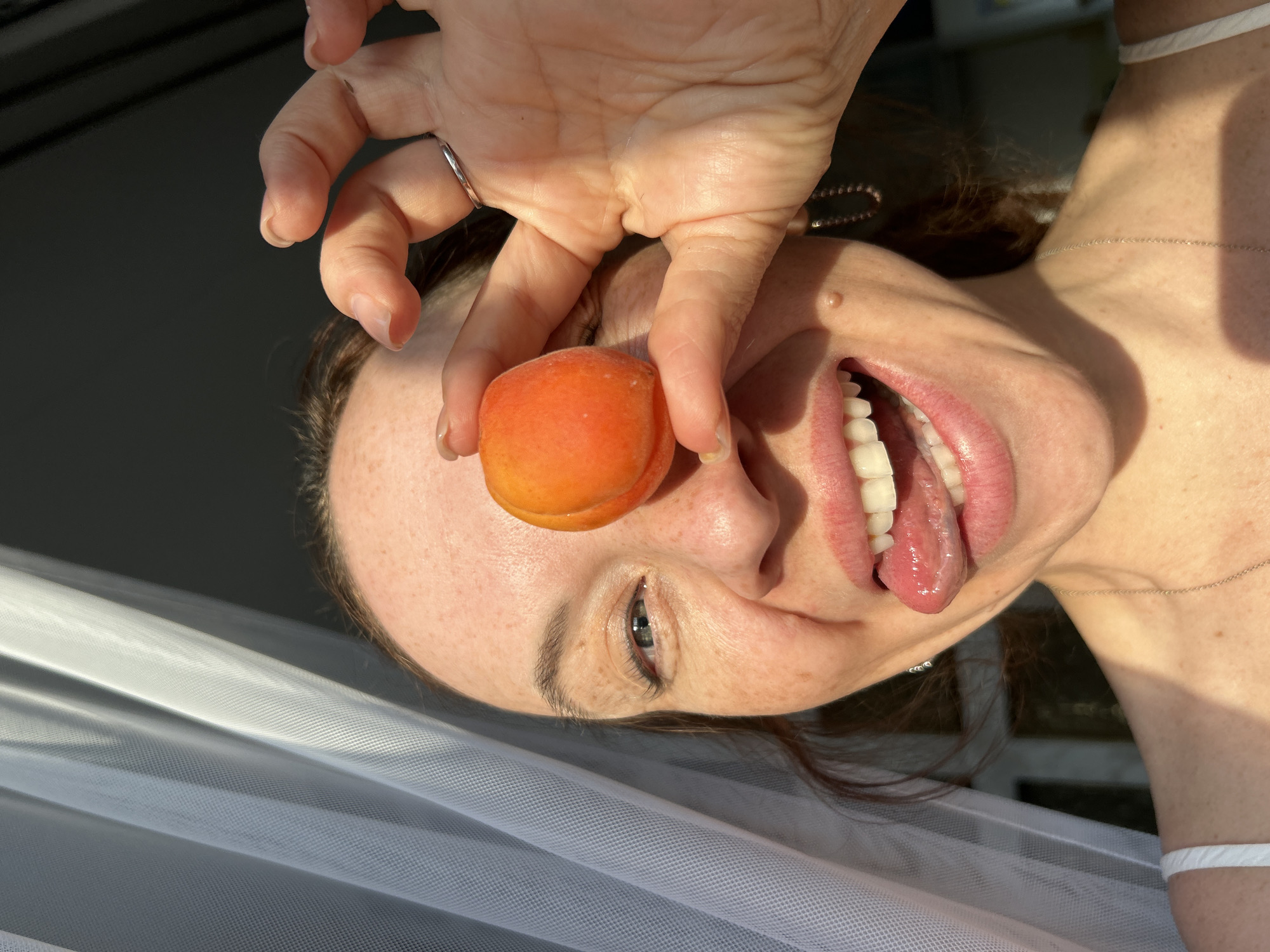A woman is holding an apricot over her eye, sticking out her tongue. She is wearing a ring on her finger and a silver necklace. In the background is a white sheer curtain, with a dark grey background visible through the window. The image was taken with a slight upward angle. There is no definitive evidence to confirm the location is in the city of Chaikovsky, Russia.