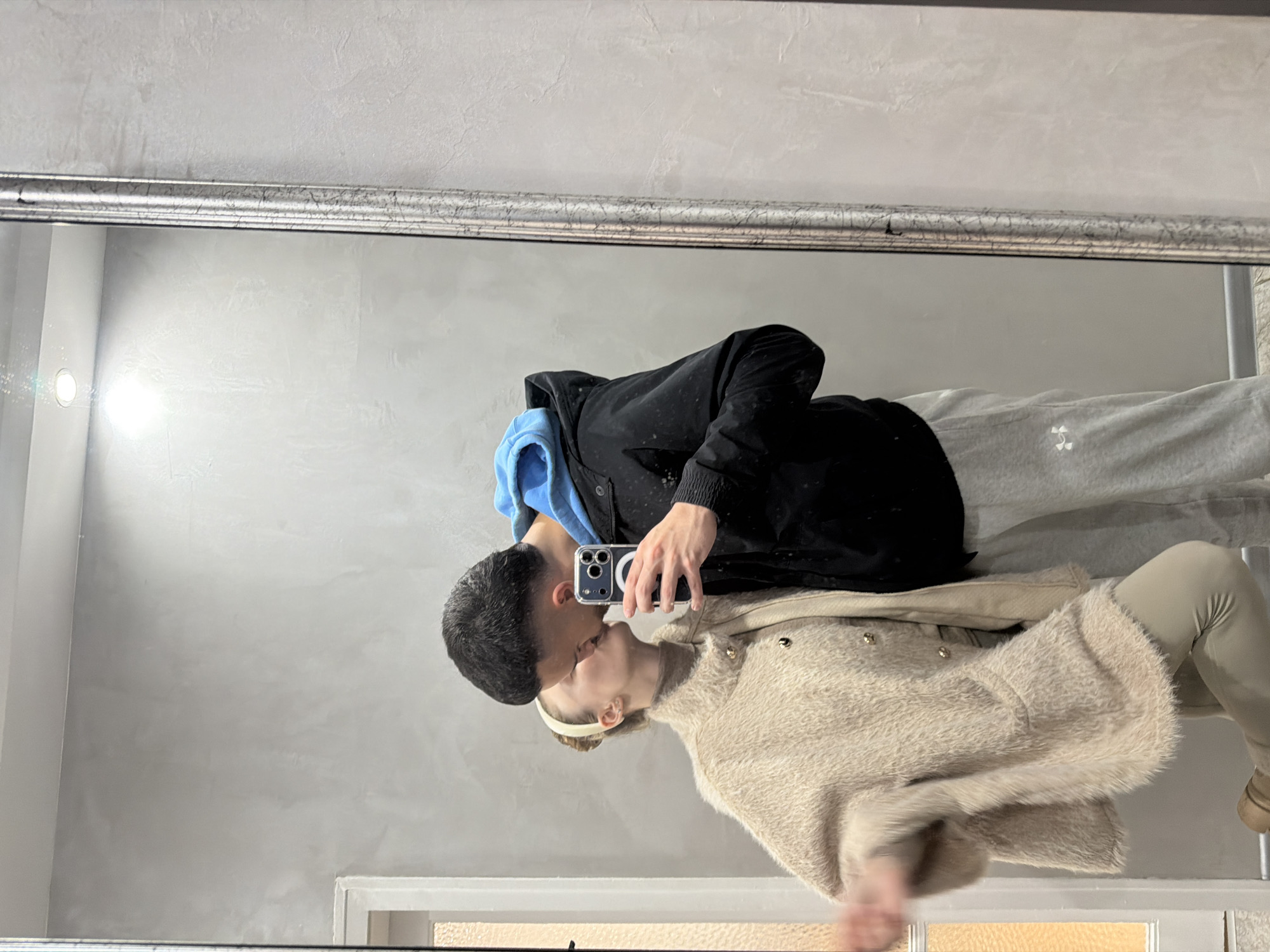 A reflection in a mirror shows two people kissing. One person wears a black jacket over a blue hoodie and light gray sweatpants. The other person wears a fuzzy beige coat and leggings. A hand holds a cell phone, presumably taking the picture. The mirror hangs on a light gray wall. The location is likely not Plovdiv, Bulgaria, given the lack of evidence in the image.