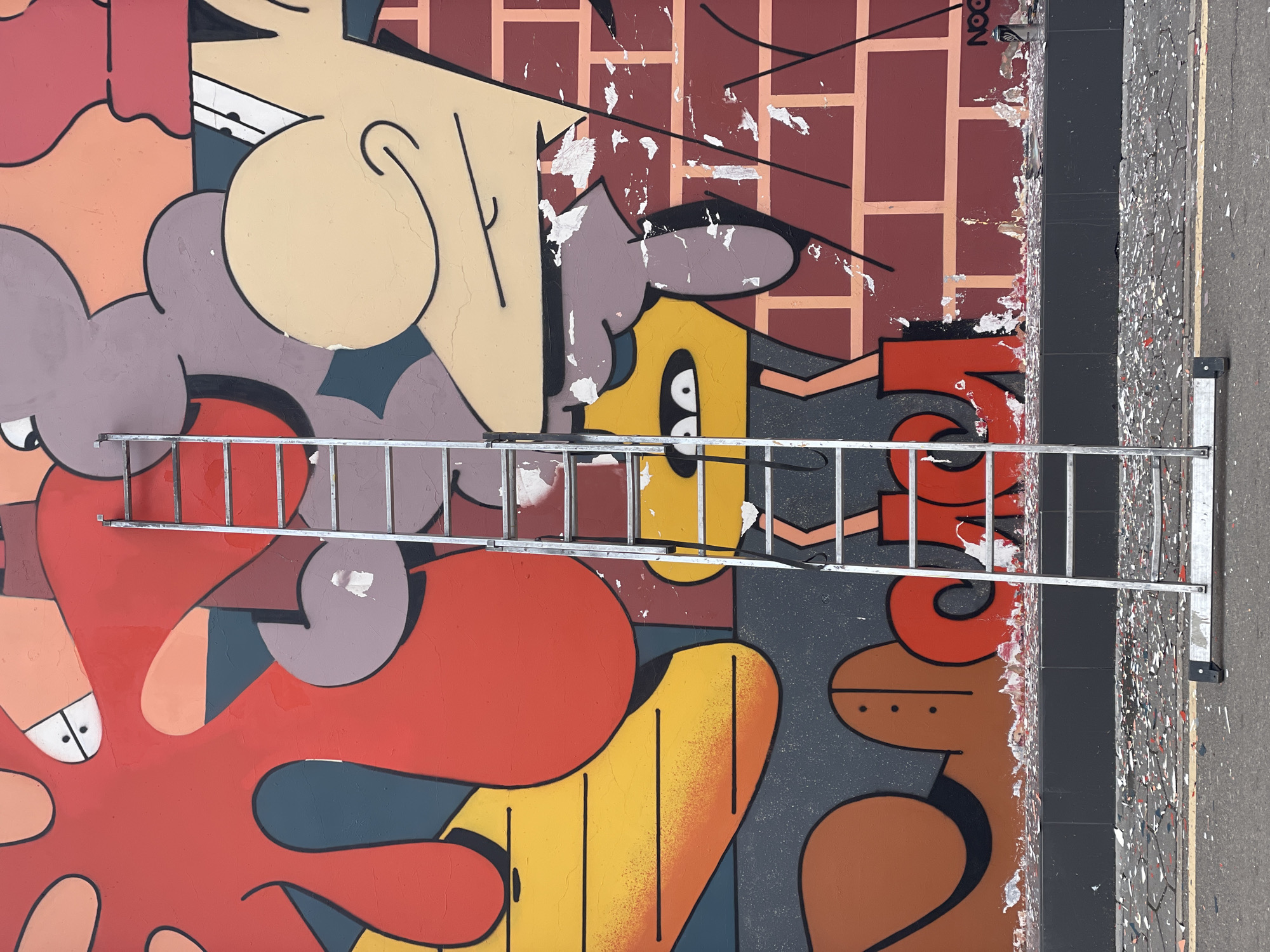 A flat, eye-level shot depicts an exterior wall with a colorful mural in Khimki, Russia. The mural consists of cartoonish figures in shades of red, yellow, orange, brown, and purple outlined with black lines. A silver ladder rests horizontally against the wall, spanning across the mural. The wall is peeling near the top, revealing the brick underneath. To the right of the wall is a narrow black curb, beyond which lies a mottled gray surface, possibly a street or sidewalk.