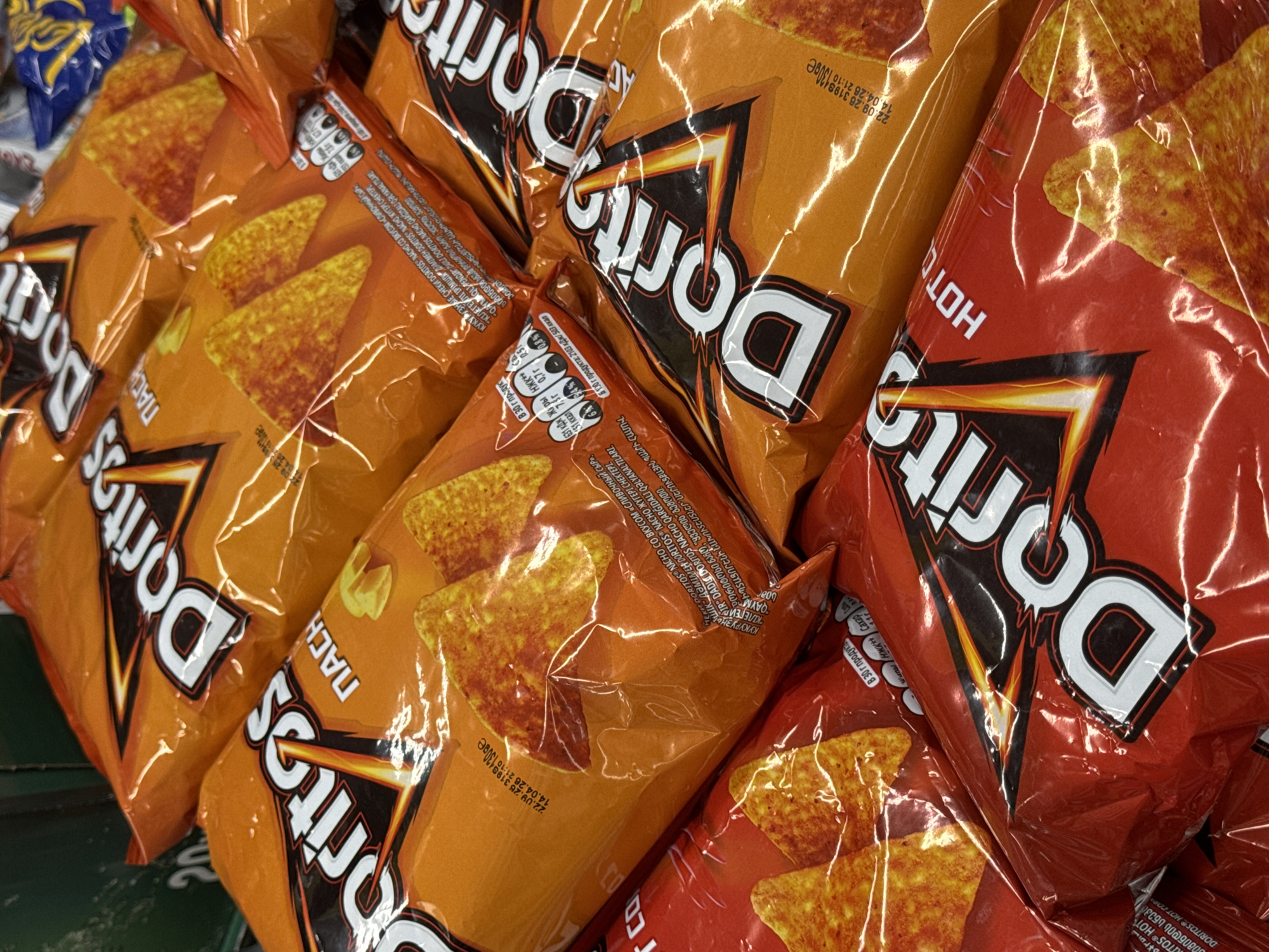The image shows a close-up of several bags of Doritos chips in a retail setting. The chips are of various flavors, including Nacho and Hot Corn, indicated by the text on the packaging. The bags are predominantly orange and red, with the iconic Doritos logo prominently displayed. The lighting suggests an indoor environment, possibly a supermarket aisle, with reflections visible on the plastic packaging. The background is out of focus, but appears to be shelves or other products. There are no people visible in the image. The scene depicts a retail display of snack food products. The location is identified as Batumi, Georgia, based on metadata associated with the image, but this information is not visually discernible from the image itself.