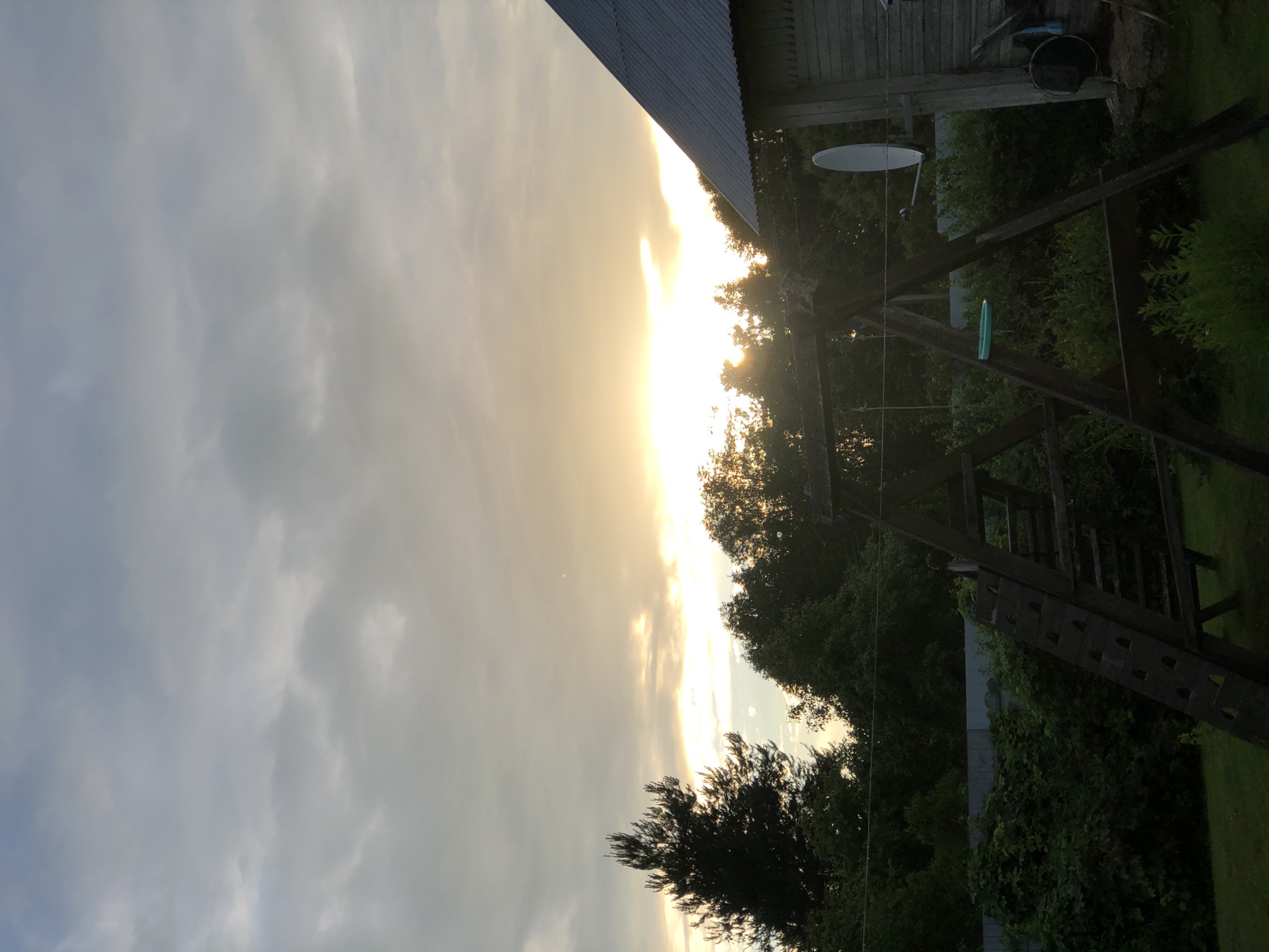 The image shows a backyard scene at dusk. The sky is filled with overcast clouds, with a bright band of sunlight peeking through the clouds on the horizon. In the foreground, a wooden swing set is visible, along with lush green vegetation. To the right, a portion of a wooden building with a corrugated metal roof is visible, and a satellite dish is mounted on its side. A tall pine tree stands to the left, silhouetted against the sky. The overall impression is one of a rural or suburban setting in Zhukovo, Russia, at the end of the day.