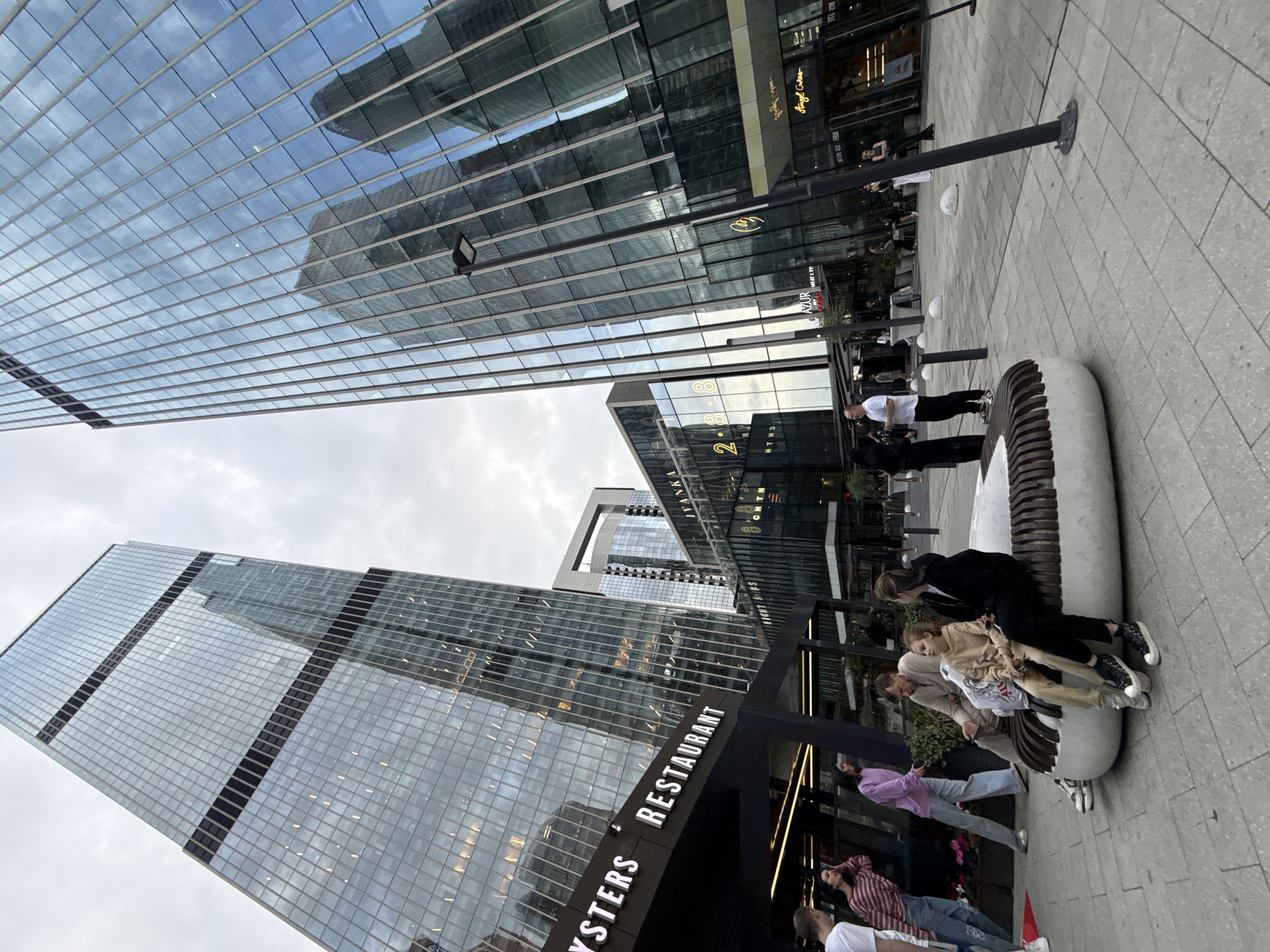 In Khimki, Russia, a wide-angle perspective captures a modern urban plaza with towering glass skyscrapers. The buildings feature reflective facades, mirroring the overcast sky and surrounding architecture. In the foreground, a family is seated on a curved, bench-like structure. A father, wearing a beige shirt and pants, sits between two daughters. The older daughter, also in beige attire, is in the center, while the younger daughter, in black, sits to his right. They appear to be conversing and are casually dressed. To the left, a man in a white t-shirt and jeans, and a woman in a striped shirt and jeans, are standing and interacting. The plaza floor is paved with grey tiles. Several shops are visible in the background, including "OYSTERS RESTAURANT," "IVANKA," and "Angel Cakes." A prominent black lamppost stands to the right of the bench. The overall scene depicts a public space with a mix of residential and commercial buildings, under a cloudy sky.