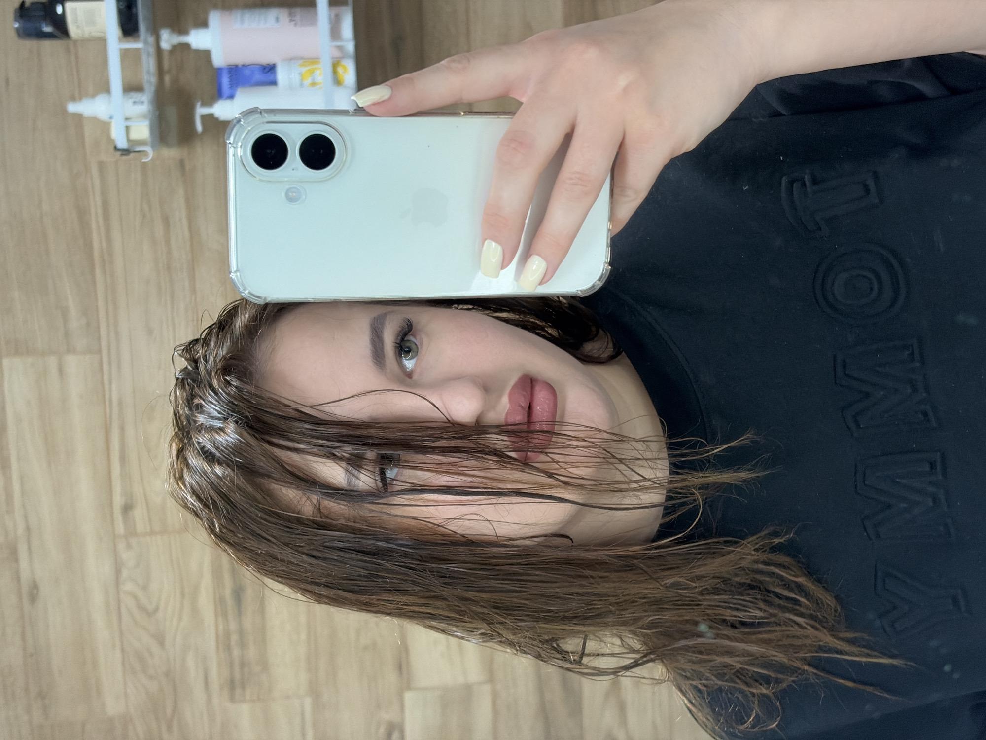 A medium shot depicts a woman holding a smartphone in her right hand, as she takes a selfie in an indoor setting. The phone, white with a clear case, obscures the top of her forehead. Her hair is wet and disheveled, partially covering her face, and appears to be light brown. Her visible skin is fair-toned, and she has dark eyes. She wears a black sweatshirt with embossed lettering on the right side. The background reveals a light brown wooden floor, and in the upper left corner, a white rack with various hair care products. The location appears to be a bathroom.