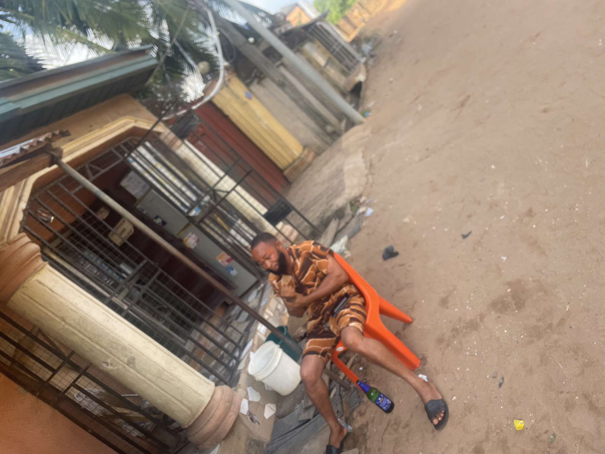 A candid, slightly tilted, medium shot captures a man sitting outdoors in what appears to be Nkpor, Nigeria. The man, with a beard and short dark hair, is seated on a bright orange plastic chair on sandy, uneven ground. He is wearing a patterned short-sleeved shirt and matching shorts in brown, orange, and white hues, along with black flip-flops. His arms are crossed over his chest, and he is smiling broadly, conveying a sense of happiness or amusement.
Near his feet, a green bottle lies on the ground, and a white plastic bucket is visible behind him. The immediate surroundings are littered with various small pieces of debris. To the left, a building with ornate, light-colored columns and dark metal security grills or gates is prominent. Palm tree fronds can be seen at the top left of the frame, suggesting a tropical climate. In the background, other structures, possibly shops or residences, with faded exteriors and rolled-down shutters, line what appears to be a street or pathway. The lighting suggests it is daytime, likely a bright morning or afternoon. No discernible text is visible.