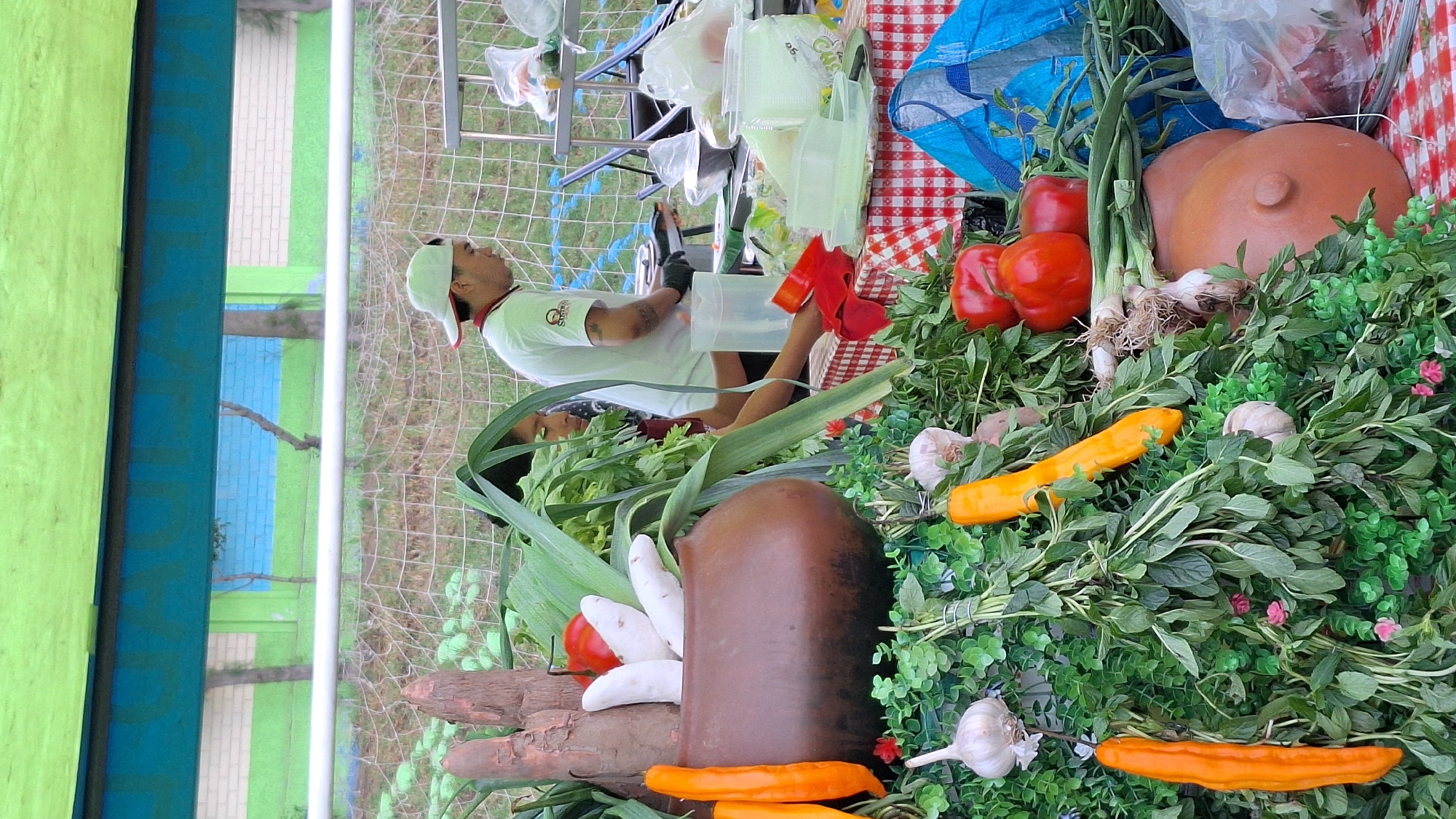 A scene in Alto Selva Alegre, Peru, features a vendor at a table. The table is covered with a red and white checkered cloth and displays a variety of produce, including red peppers, garlic, yellow peppers, green onions, and herbs. Also on the table are a blue bag and plastic containers. In the background, a person wearing a white cap and white shirt stands behind the table, appearing to handle a plastic container. A metal cart is positioned to the left of the person. Behind the vendor's table is a metal fence. Further back, a green and blue wall is visible. In the foreground, there is a decorative arrangement of wooden objects, white root vegetables, and a brown clay pot.