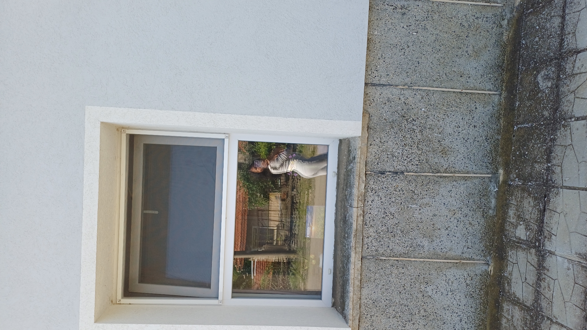 A low-angle shot depicts the corner of a building. The building's wall, featuring a rough, white texture, meets a gray, multi-textured exterior ground surface or sidewalk. A window is set into the wall; a partially open screen is visible on the left, and a reflection in the glass pane shows a person holding a camera or phone and an outdoor scene with a building with a red roof. The ground or sidewalk is made of varied materials, including patterned tiles and what appears to be poured concrete with separating strips. The location is Gorna Verenitsa, Bulgaria.