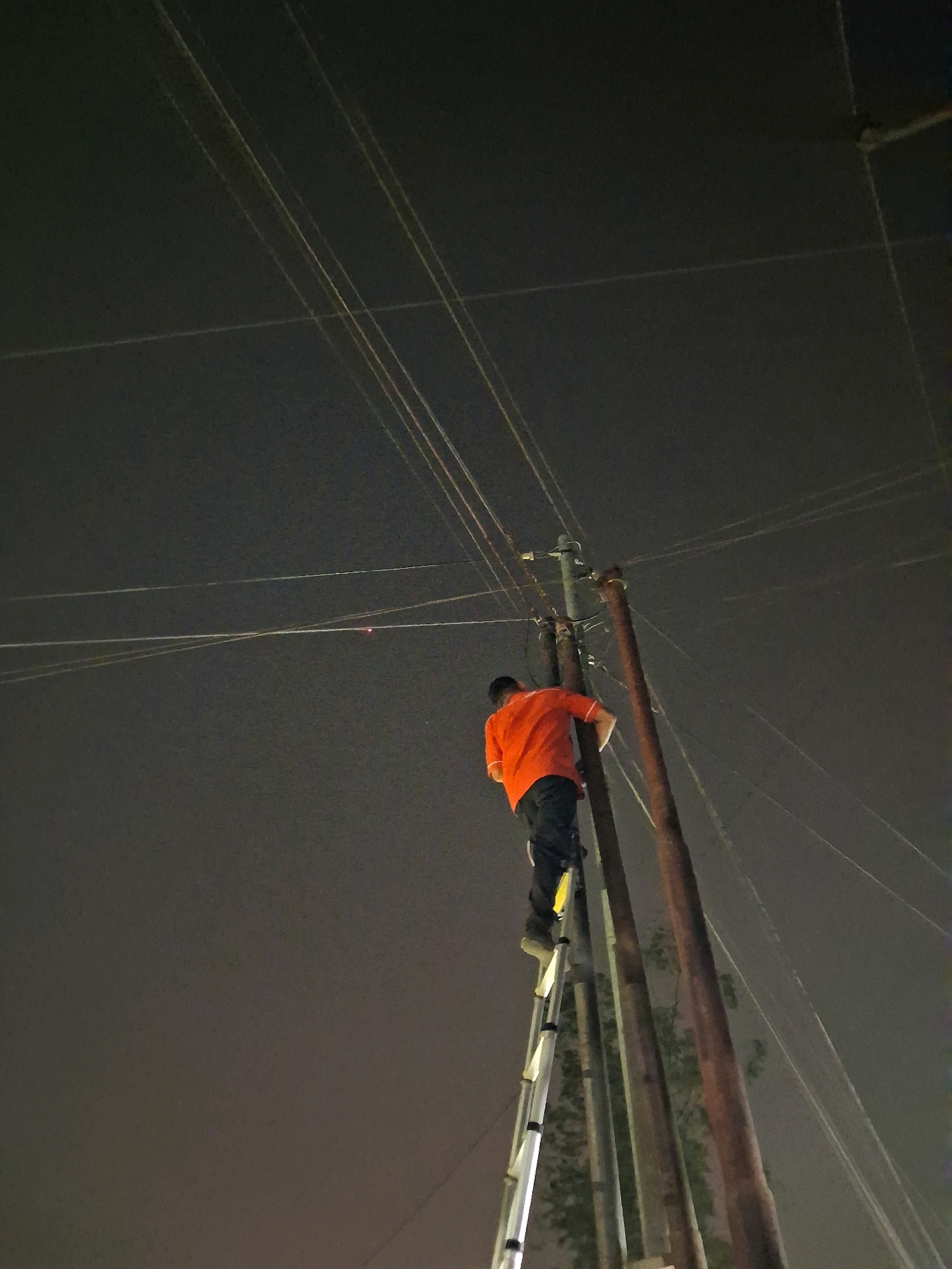 An adult male wearing an orange shirt and dark pants is seen from behind climbing a metal ladder. The ladder is positioned against a wooden utility pole. Multiple power lines are visible stretching across the dark, overcast sky. The scene appears to be in Pangkalan Kerinci, Indonesia, at night.