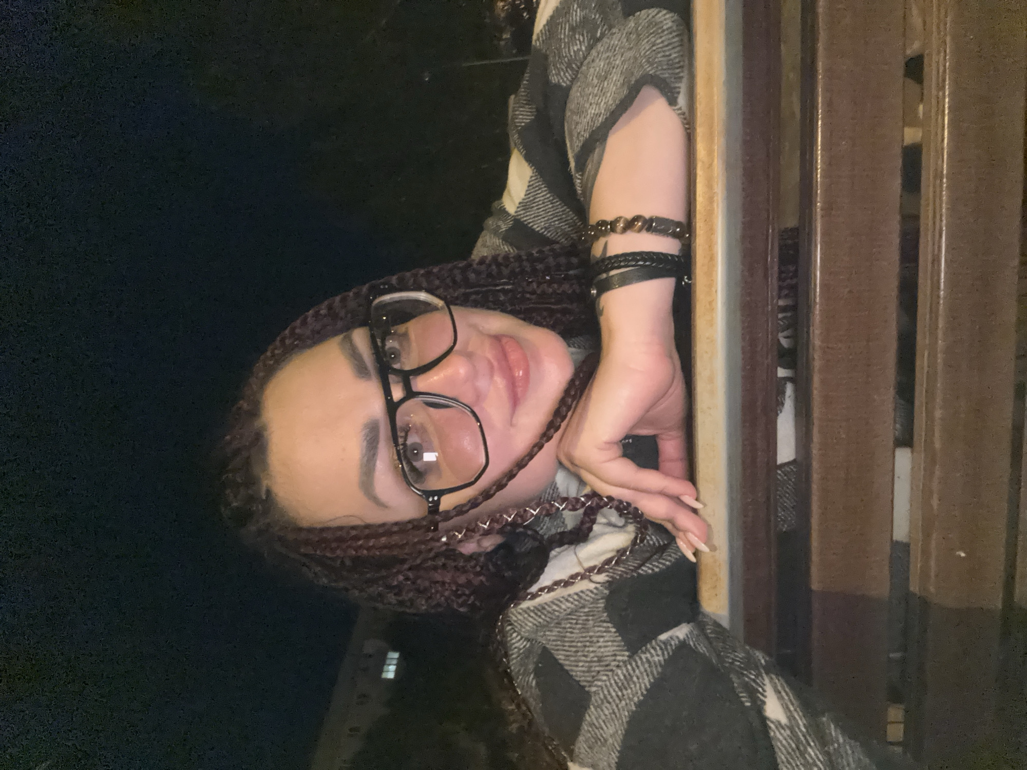 A medium shot taken at night shows a woman with braided hair and glasses leaning on a wooden surface. She is wearing a checkered jacket and bracelets on her wrist. The background is dark with some blurry lights suggesting an urban setting. The exact location in Tolyatti, Russia, is not discernible from the image.
