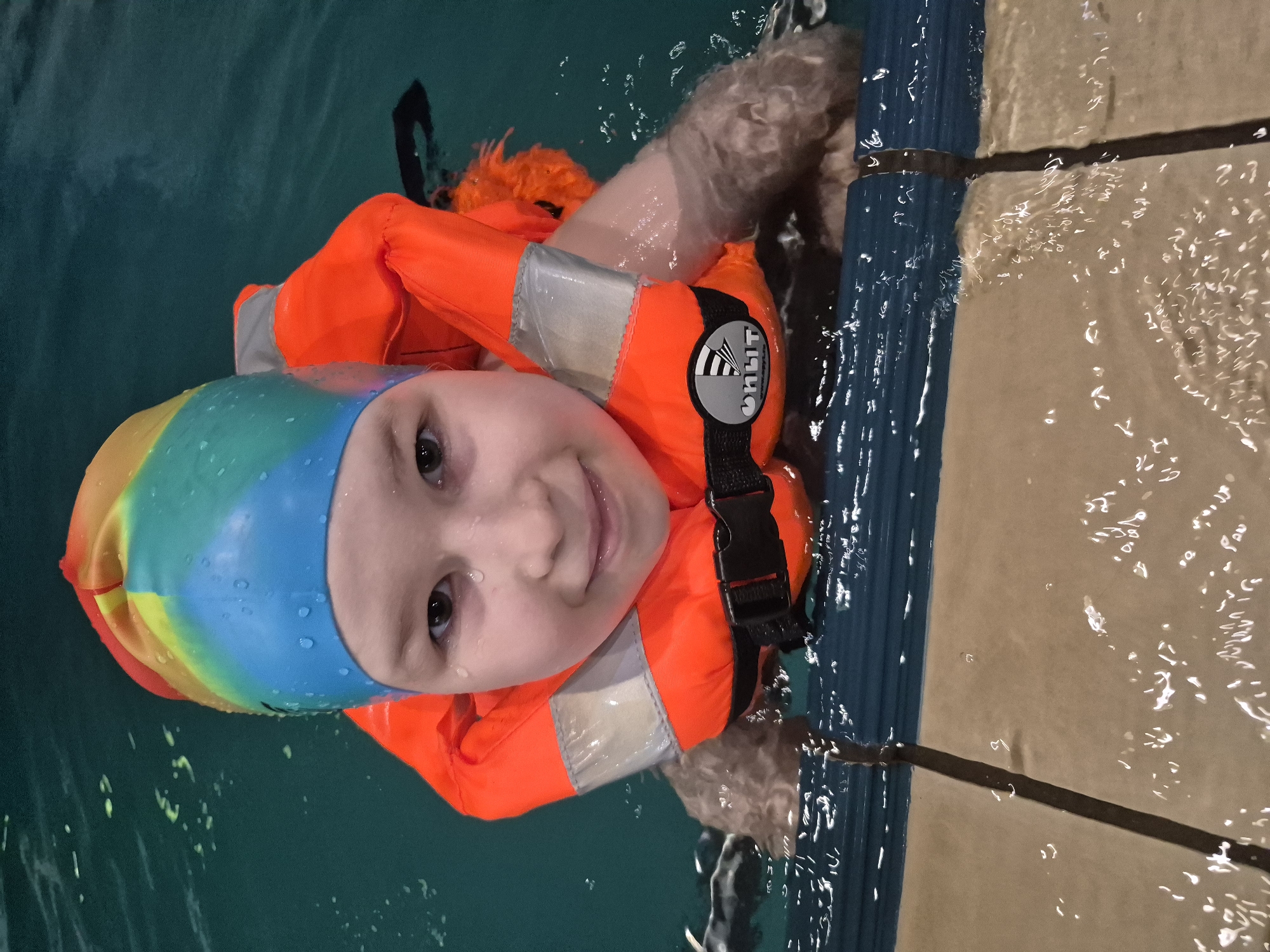 A young child is in a swimming pool, wearing a rainbow-colored swim cap and an orange life vest. The child is holding onto the side of the pool, which is bordered by a blue tile and a light-colored tile. The location of the image is the city of Tobolsk, Russia.