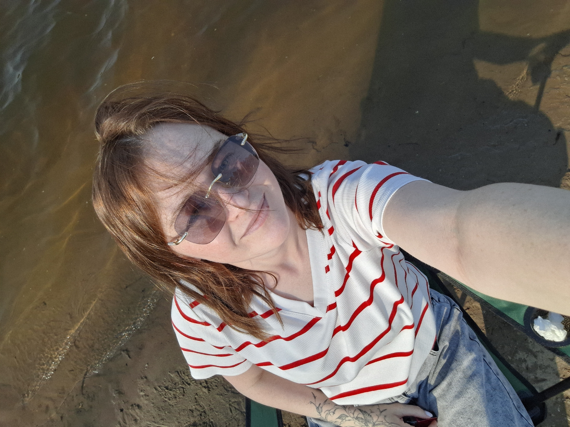 A woman is taking a selfie while sitting in a folding chair near a body of water, possibly on a bank in the city of Chaikovsky, Russia. She wears sunglasses, a white, red-striped shirt, and light blue jeans. A tattoo is visible on her left forearm. The water appears brownish and calm, and a portion of the sandy bank is visible. The shadow of a person is cast on the shoreline.