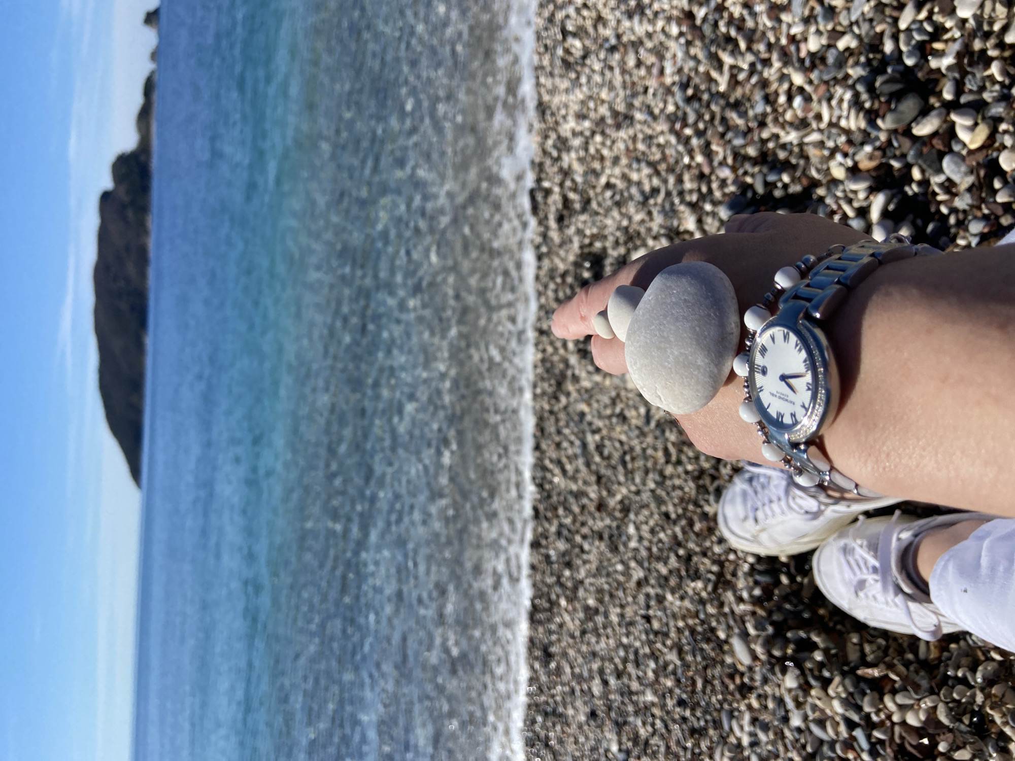 The image captures a scene on a pebble beach in Budva, Montenegro, likely along the Adriatic Sea. The view is from an elevated, first-person perspective. In the foreground, a person's hand is visible, adorned with a silver watch with a white face and a decorative beaded bracelet. A large, smooth, white pebble is held between the thumb and index finger, as if displaying it. The person is wearing white canvas shoes and white trousers.
The middle ground features the shoreline where the waves gently wash onto the pebble beach. The pebbles are a mix of gray, brown, and black.
The background depicts the blue-green sea and a rocky cliff formation on the left side of the frame. The sky is a clear, light blue.