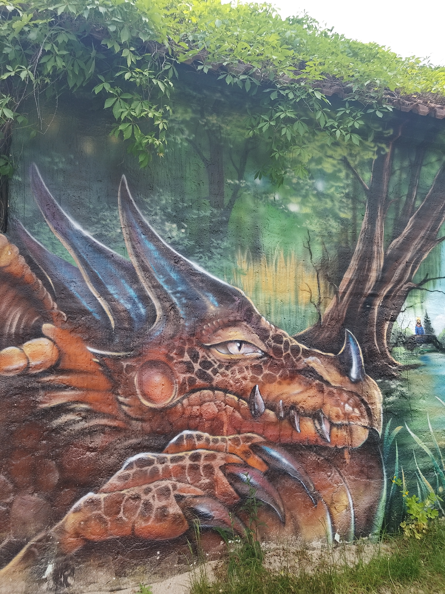 A detailed, vibrant mural on a textured concrete wall depicts the head and clawed foot of a dragon. The dragon features brown, orange, and blue coloring with sharp horns and teeth. The wall behind the dragon is painted with a green forest scene and a small body of water. To the right of the dragon, near the edge of the water, is a small figure of a man with a beard. Live green vines and leaves cover the top of the wall, partially obscuring the painted forest. Patches of grass and weeds grow along the base of the wall in the foreground. The location is in the city of Unknown, Unknown.