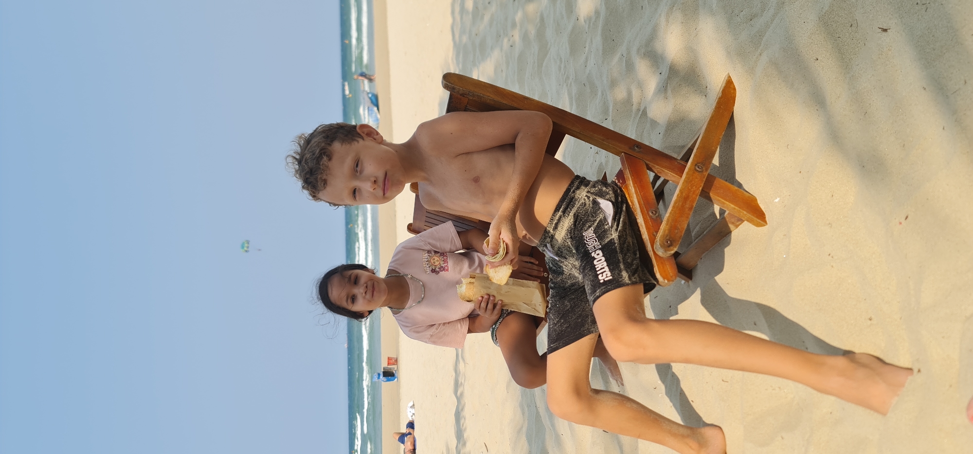 The image captures two children enjoying a sunny day on a beach in Đà Nẵng, Vietnam. In the foreground, a boy with curly brown hair, shirtless and wearing black swim shorts adorned with the white text "TUSH SPORTS!", sits on a wooden beach chair. He looks towards the camera with a subtle expression. Beside him, a girl with dark hair, dressed in a light pink t-shirt featuring a sun graphic and a silver necklace, smiles at the camera while holding what appears to be food, possibly a sandwich, wrapped in paper. Both children seem relaxed, suggesting they are having a snack or meal on the beach.
The setting is a vast sandy beach, stretching towards a clear blue ocean with gentle waves. In the middle ground, other beachgoers can be seen enjoying the water and shore. The sky is bright and cloudless, and in the distance, a parasailor is visible over the ocean, indicating popular water sports activities. The bright sunlight casts distinct shadows on the sand from the beach chair, suggesting it is a clear day, likely late morning or afternoon. The overall scene depicts a pleasant and typical beach vacation moment.