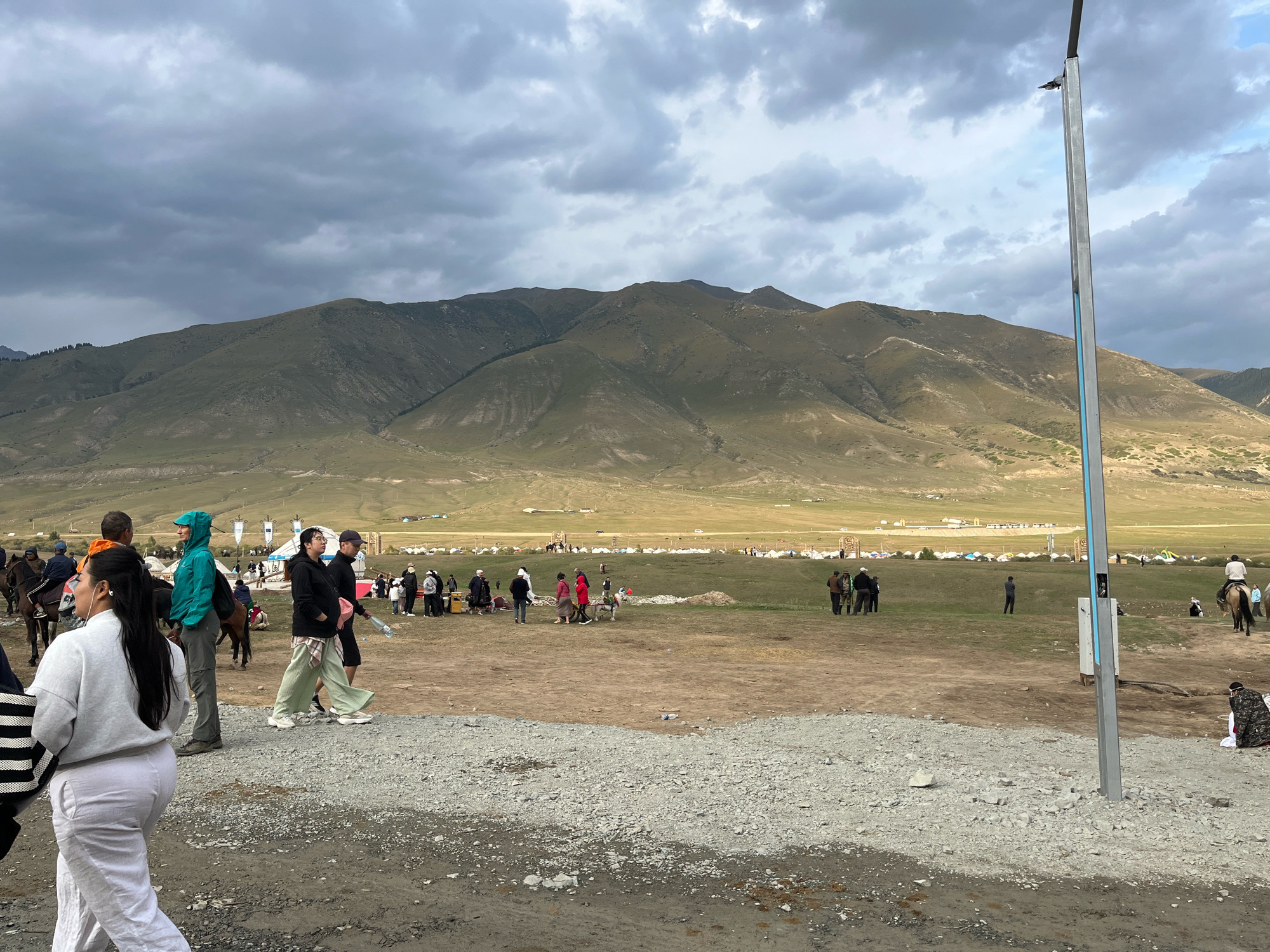 The image depicts an outdoor gathering, likely a festival or encampment, with a vast mountain range serving as a backdrop under a cloudy sky. In the foreground, several individuals are walking on a gravelly path. To the left, a person in a white sweatshirt and pants walks away from the camera, carrying a striped bag. Further back, a group of people are seen in various interactions: some are conversing, others are walking, and a few are mounted on horses. Tents are visible spread across the grassy field in the mid-ground, indicating a temporary settlement. The overall environment is open and rural, with a tall, grey utility pole positioned in the right foreground, partially obscuring the view of the distant tents and people. This scene appears to be set in the vicinity of Semenovka, Kyrgyzstan, given the mountainous terrain.